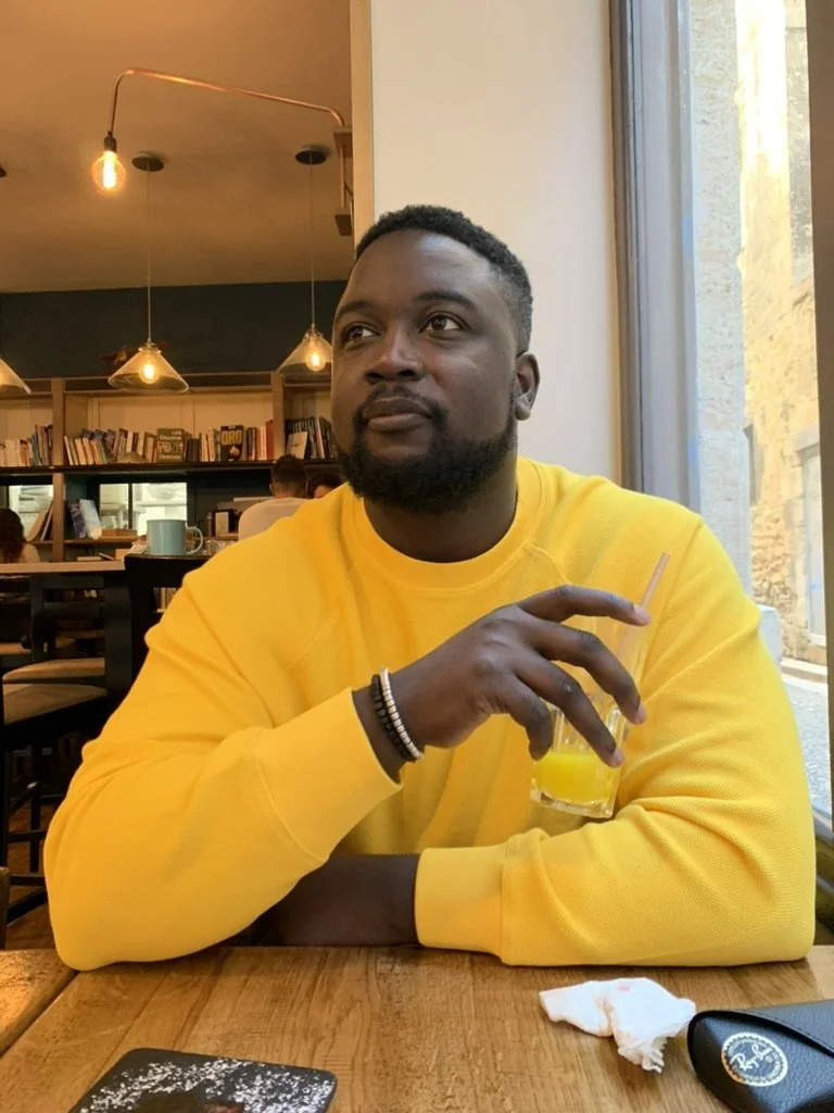 Un homme assis dans un café, portant un sweat jaune, tenant un verre de jus d'orange, avec une bibliothèque en arrière-plan et un éclairage chaud.