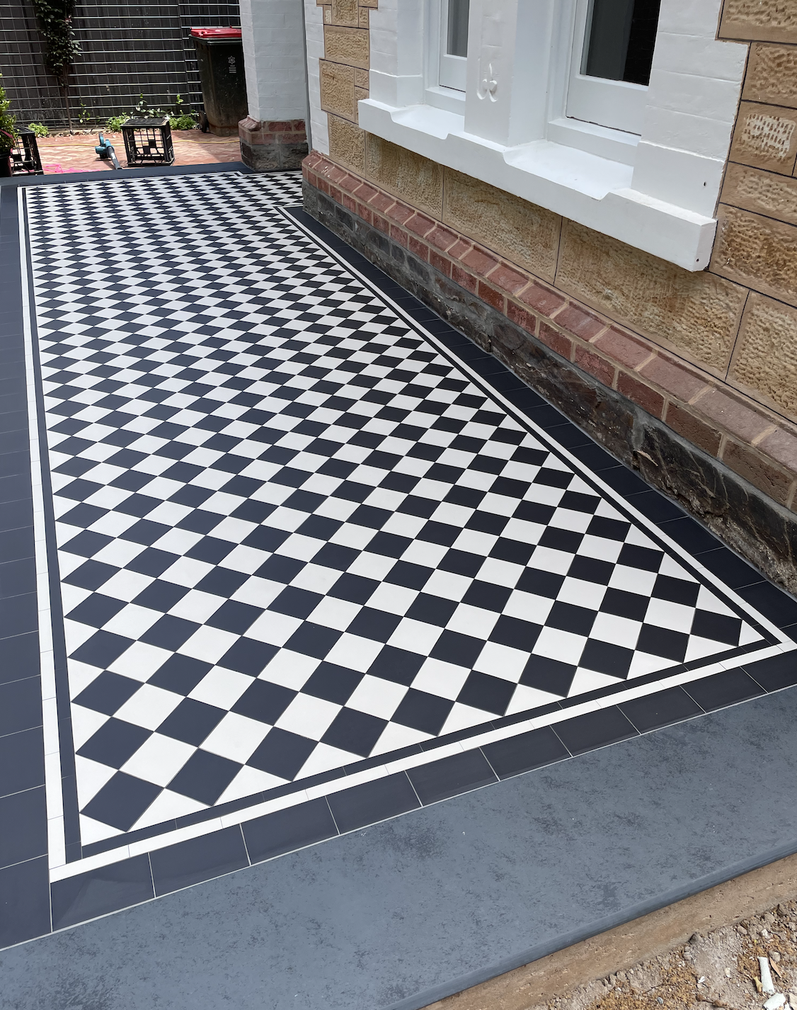 A checkered black and white tile patio outside a house, bordered by black tiles and a gray concrete edge, with a brick and stone house wall in the background.