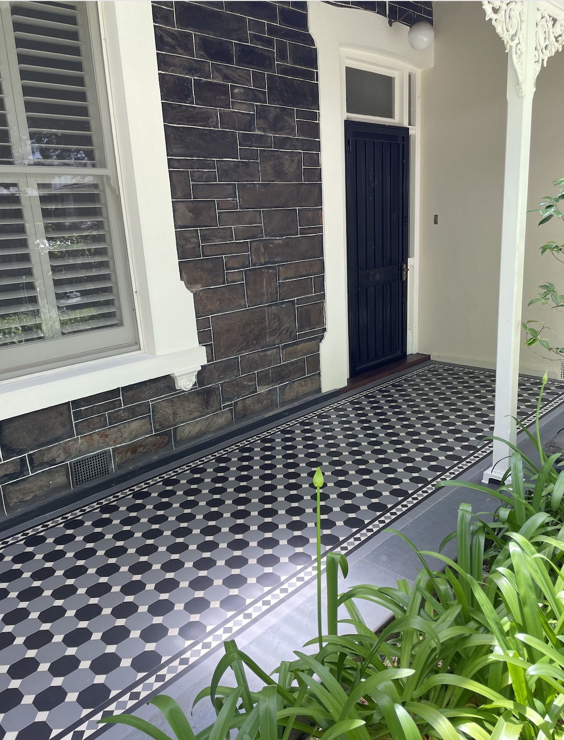Porch area with patterned black and white tile flooring, window with blinds, dark front door, and decorative white trim. Green plants are in the foreground.