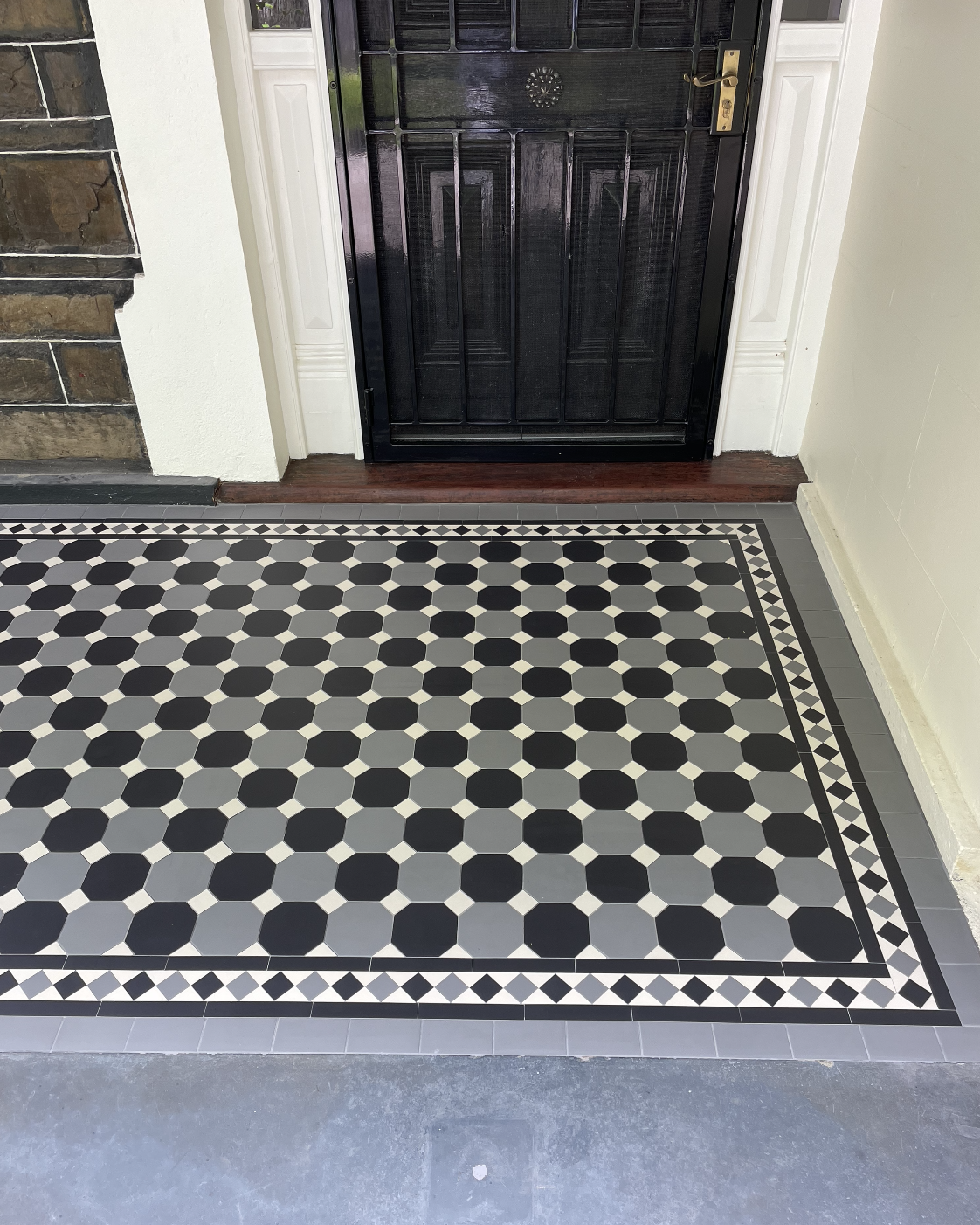 A patterned black, gray, and white tiled floor in front of a black door with a gold handle.
