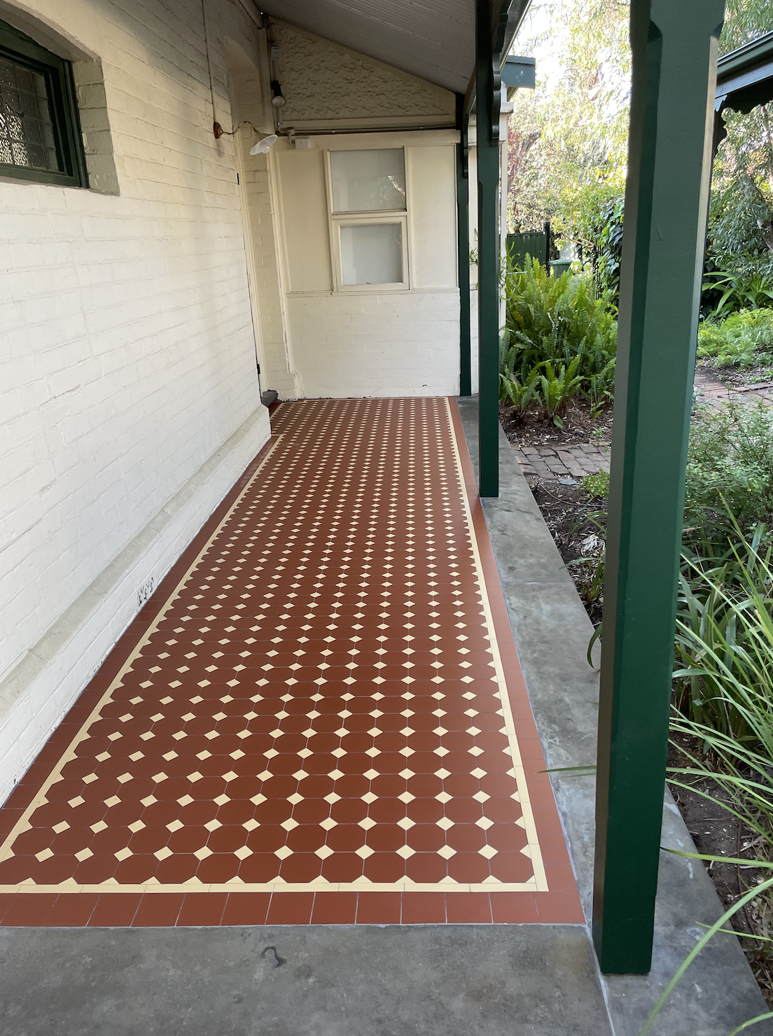 Porch with a decorative red and cream tile floor, white brick wall, green posts, and a small garden area with green plants beside it.