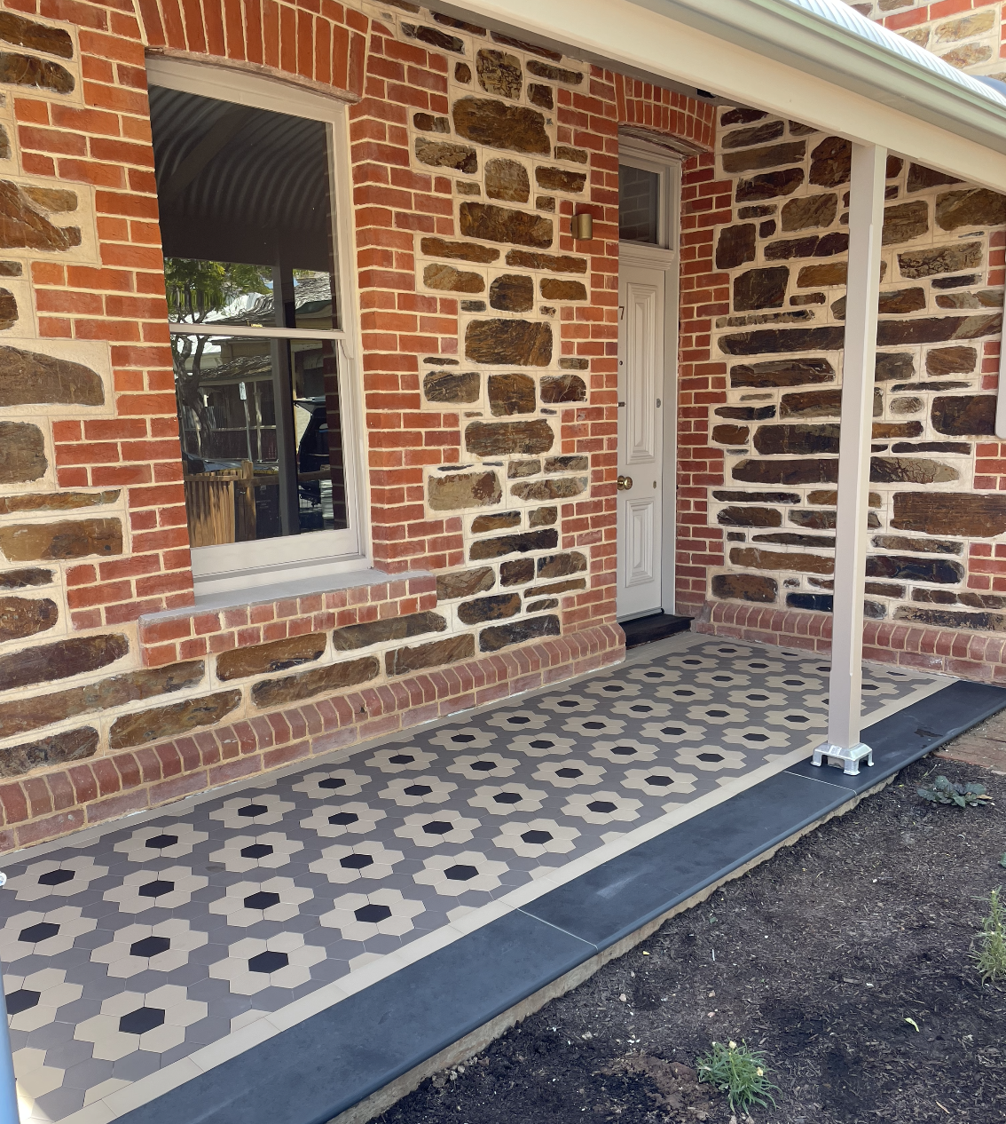 The front porch of a brick house with patterned tile flooring, a white door, and a window. The porch has a metal support post and a small area of soil with a few plants.