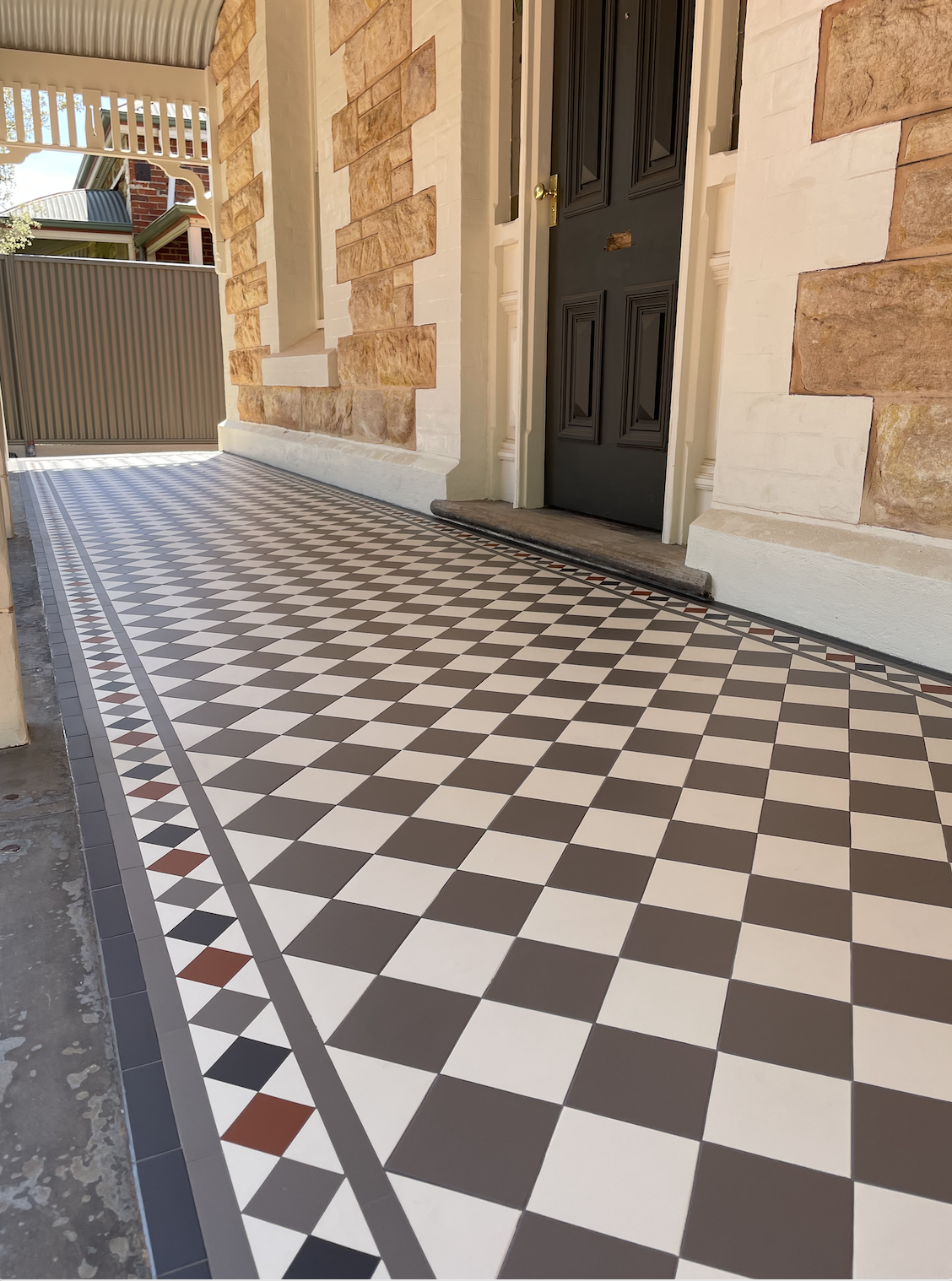 Front porch with patterned tile flooring, black front door with mail slot, brick and white wall, and a black metal gate in the background.