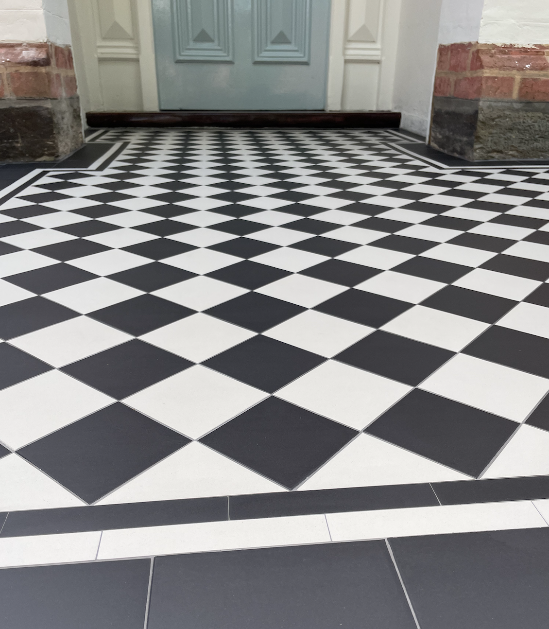 Black and white checkered tile floor leading to a closed light blue door with white trim and brick walls on either side.
