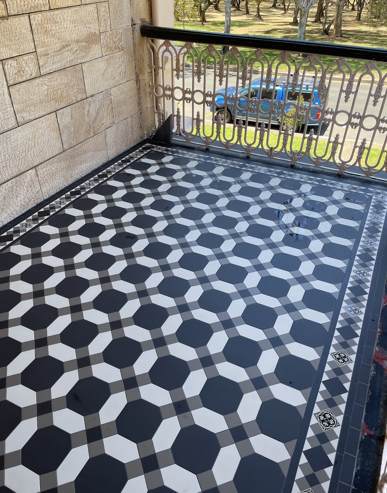 View of a tiled balcony with black, white, and gray geometric pattern, a decorative metal railing, and a parking lot with a blue vehicle and trees in the background.