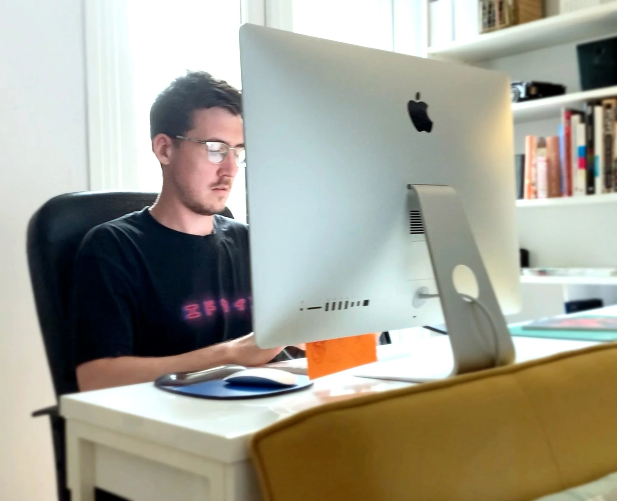 A man with glasses working at a white desk on an Apple iMac computer, with a bookshelf in the background.