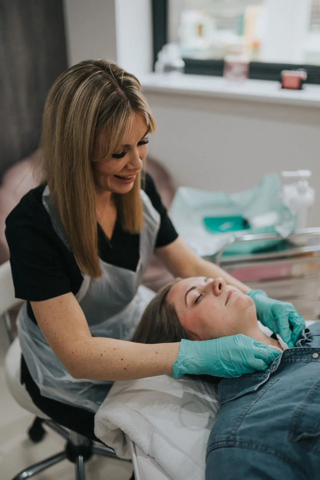 A woman with blonde hair and a black shirt receives a facial treatment from a skincare professional wearing gloves, while lying on a treatment bed in a clinical setting.