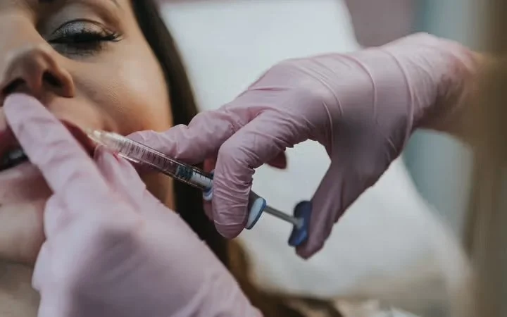A woman receiving a facial injection with a syringe while lying on a treatment bed, wearing pink gloves.