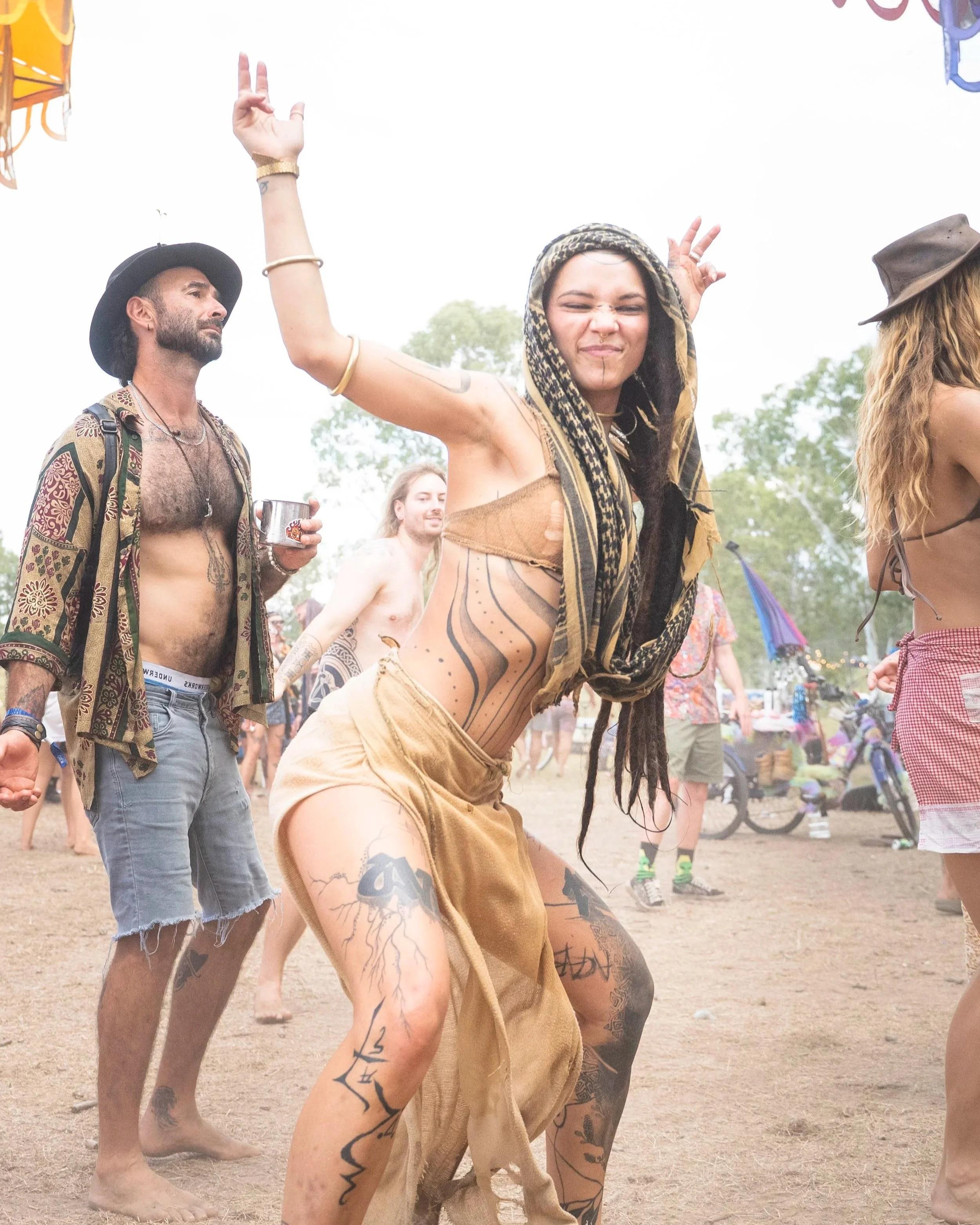People dancing at an outdoor music festival, woman with tattoos and headscarf making a peace sign, man with hat and tattoos holding a drink, others in casual festival attire, festival tents in background