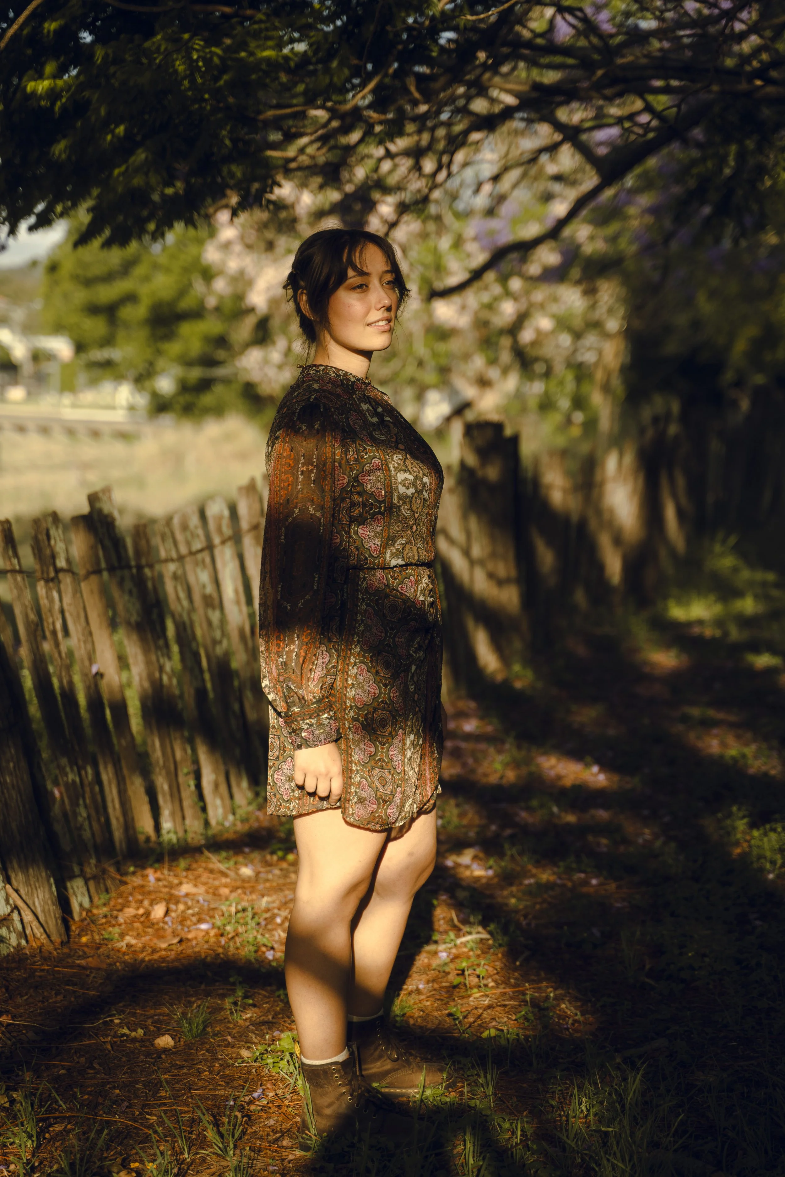 A woman with dark, wavy hair standing outdoors in warm sunlight, wearing a long-sleeved, patterned dress and brown boots, near a rustic wooden fence under a tree with pink blossoms.