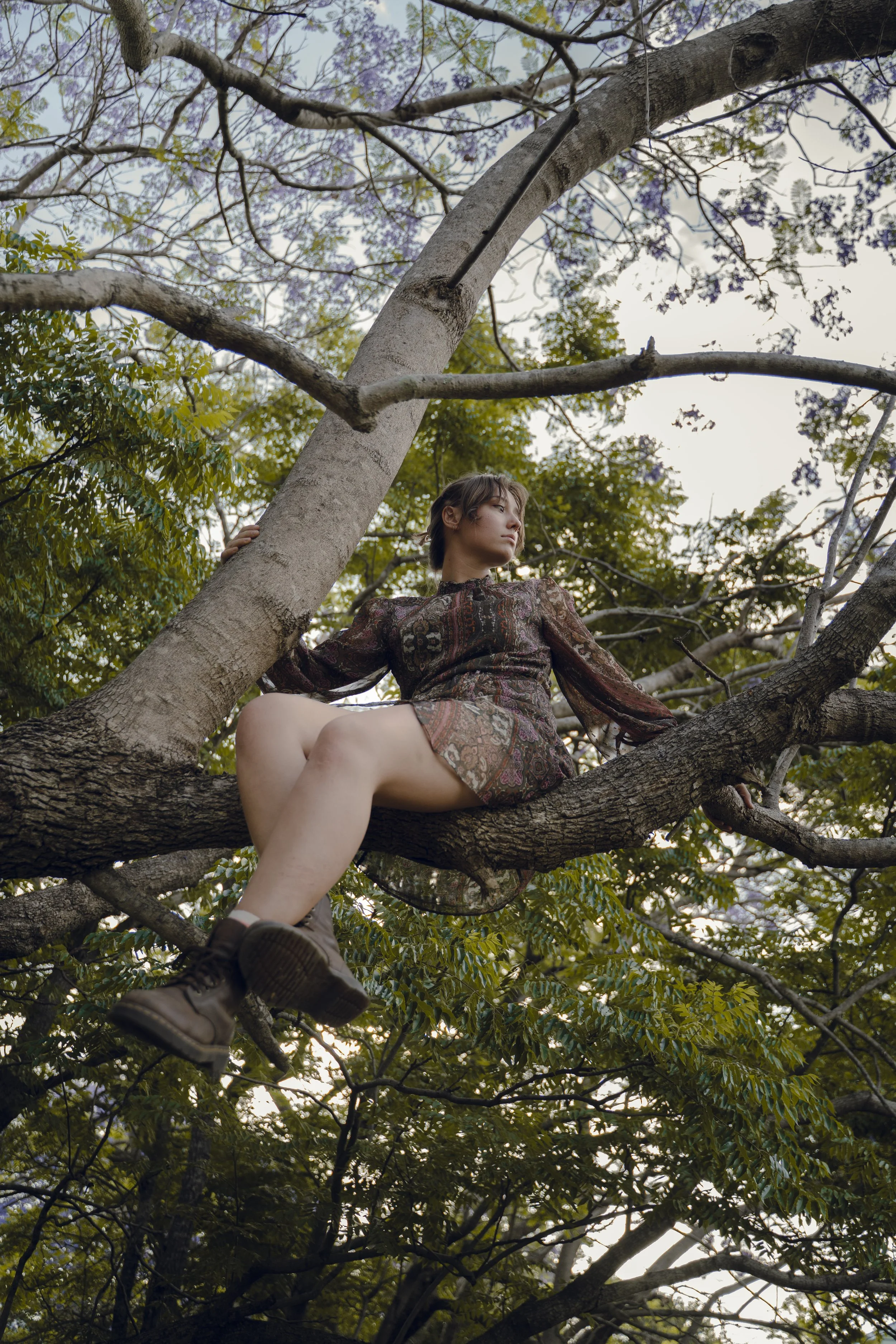 A young woman sitting on a tree branch surrounded by green leaves and purple flowers, wearing a patterned dress and brown boots.