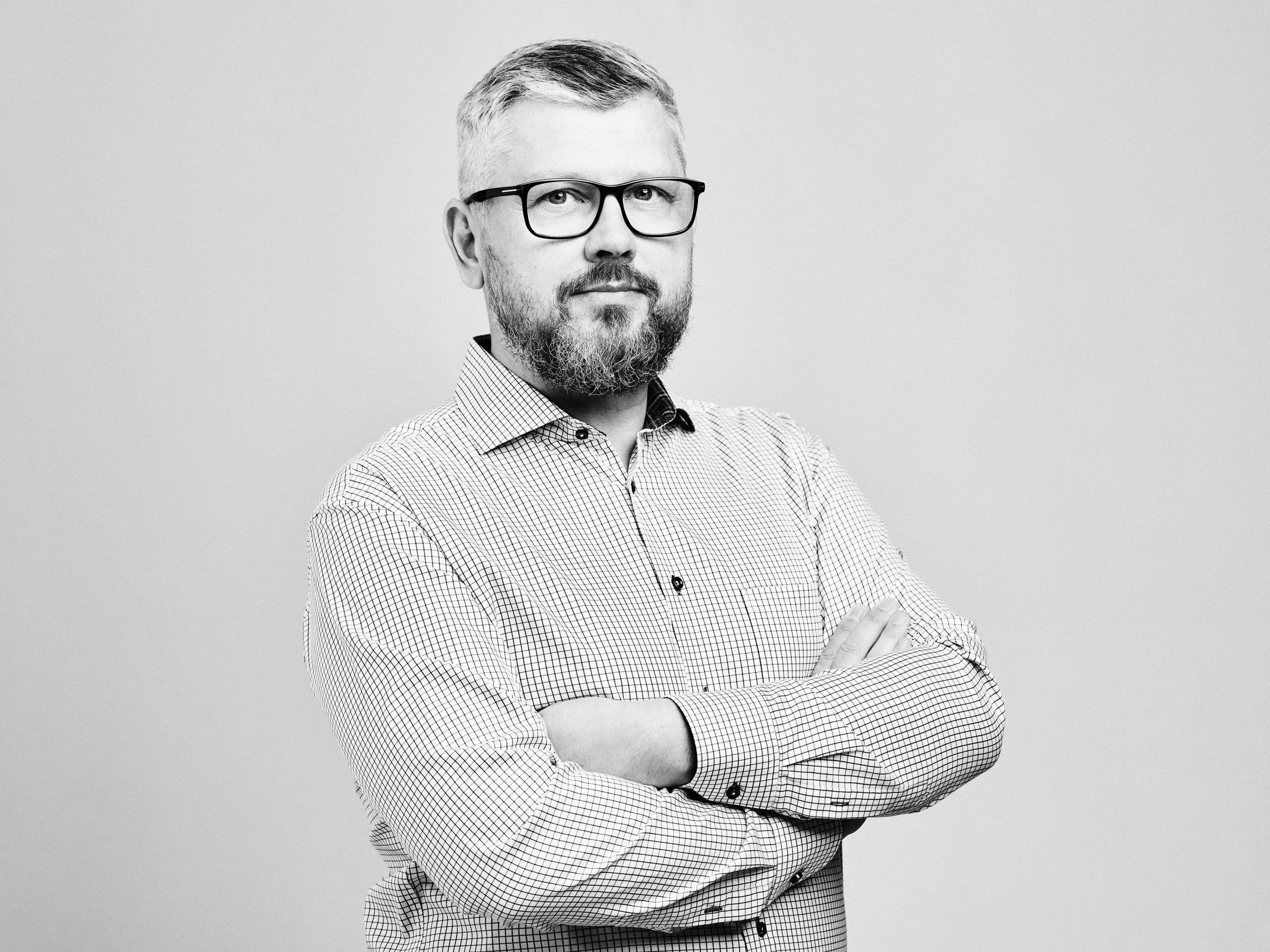 A black and white portrait of a middle-aged man with glasses, a beard, and short hair, wearing a checkered button-up shirt with arms crossed against a plain background.