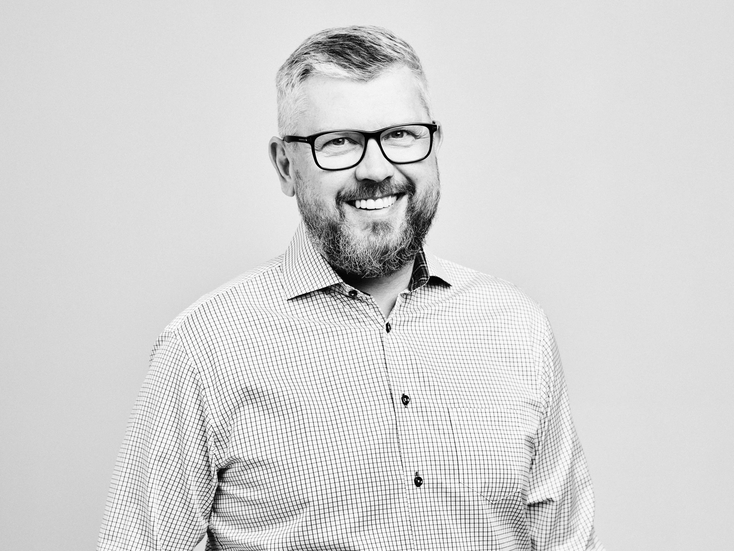 Black and white portrait of a man with glasses, short hair, a beard, and a checkered shirt, smiling against a plain background.