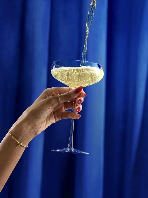 A hand holding a champagne glass as golden bubbly liquid is being poured into it against a blue curtain background.