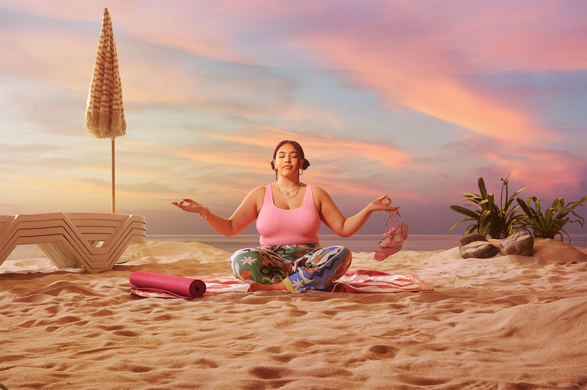A woman practicing yoga on a sandy beach during sunset, sitting cross-legged on a striped towel, with her eyes closed, wearing a pink top and colorful pants, holding a sandal in one hand and the other hand in a mudra position, with a rolled-up yoga mat nearby and plants on the right side.