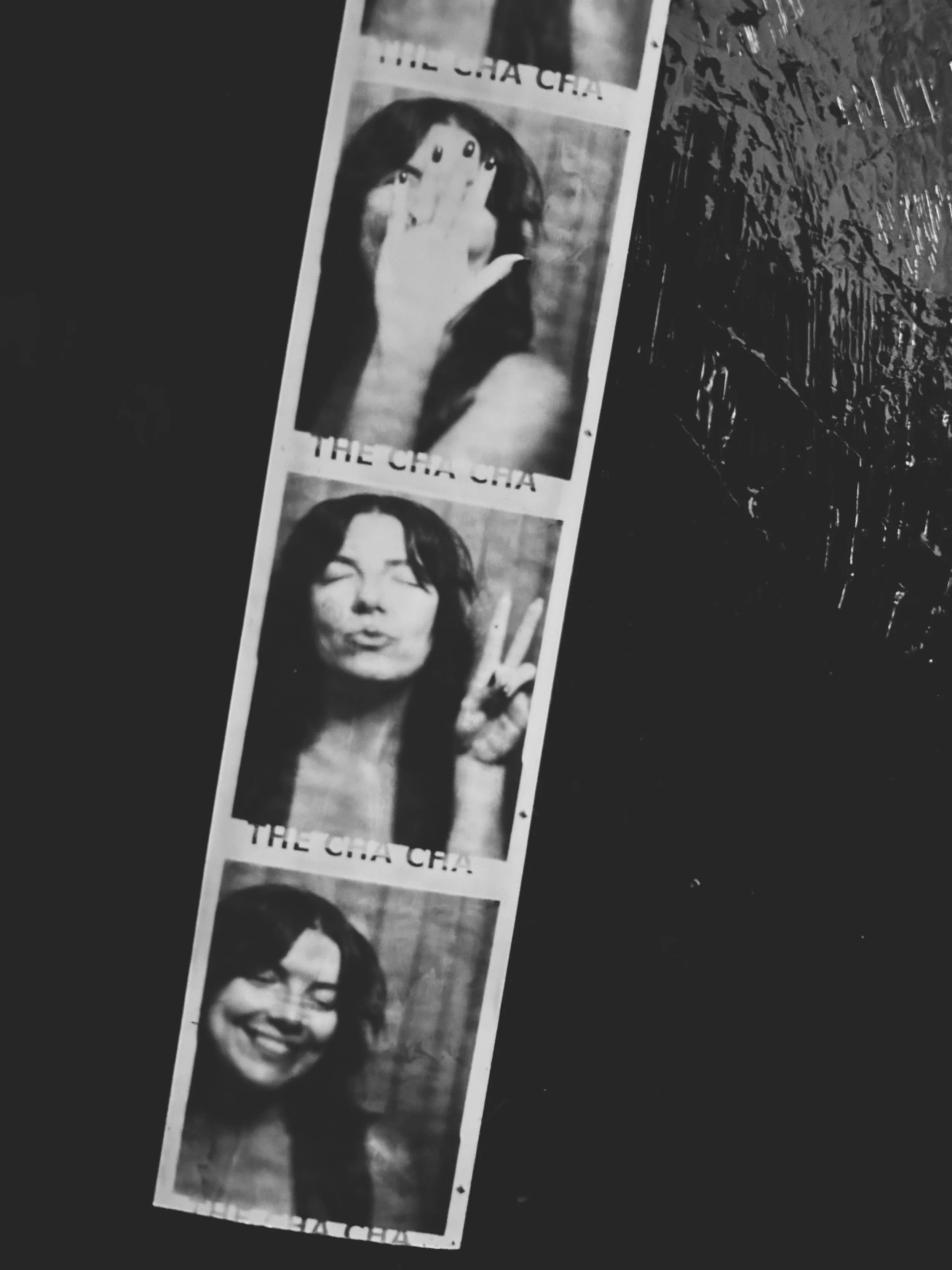 A strip of three black and white photo booth pictures of a woman making different facial expressions and hand gestures, with text partially visible at the top and bottom of the strip.