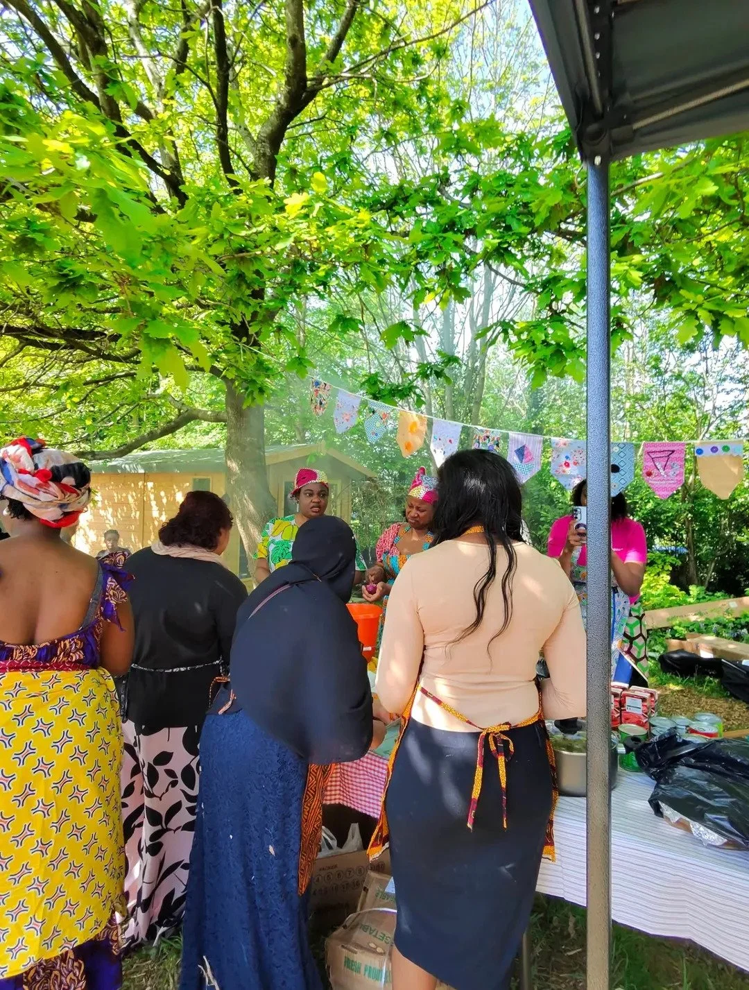 Group of women gathered outdoors under a large leafy tree, at a daytime social event with colorful bunting hanging in the background, some women wearing traditional African dresses and headwraps.