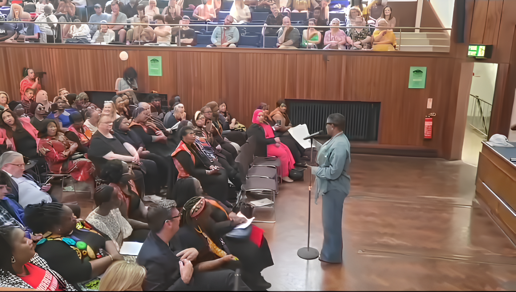 A speaker in gray suit and glasses stands at a microphone addressing a seated audience in a wood-paneled auditorium with a balcony. The audience includes diverse women, some with headscarves, some taking notes, and some using phones.