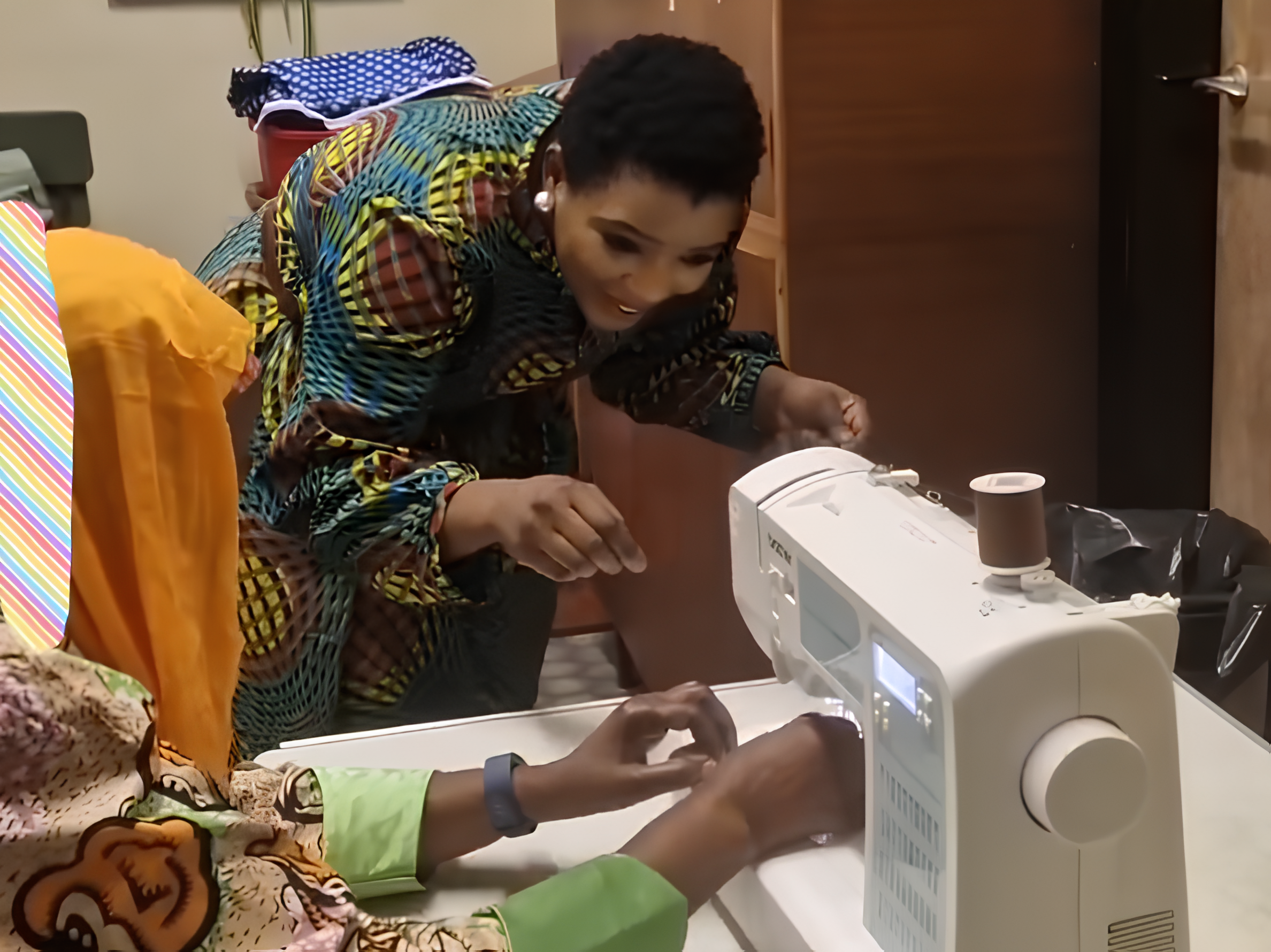 A woman in colorful clothing looking at a sewing machine while another person, possibly a woman, is smiling and wearing colorful attire at the sewing machine.