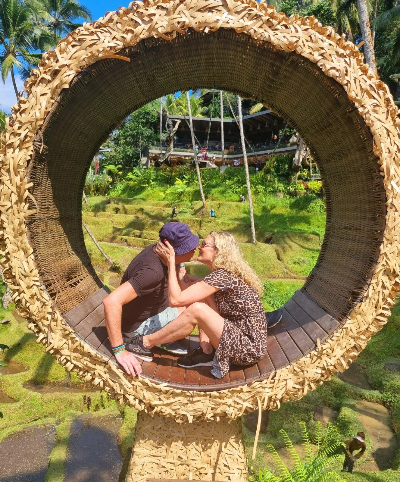 Een stel zit in een grote, ronde, gevlochten rieten sculptuur en leunt naar elkaar toe.