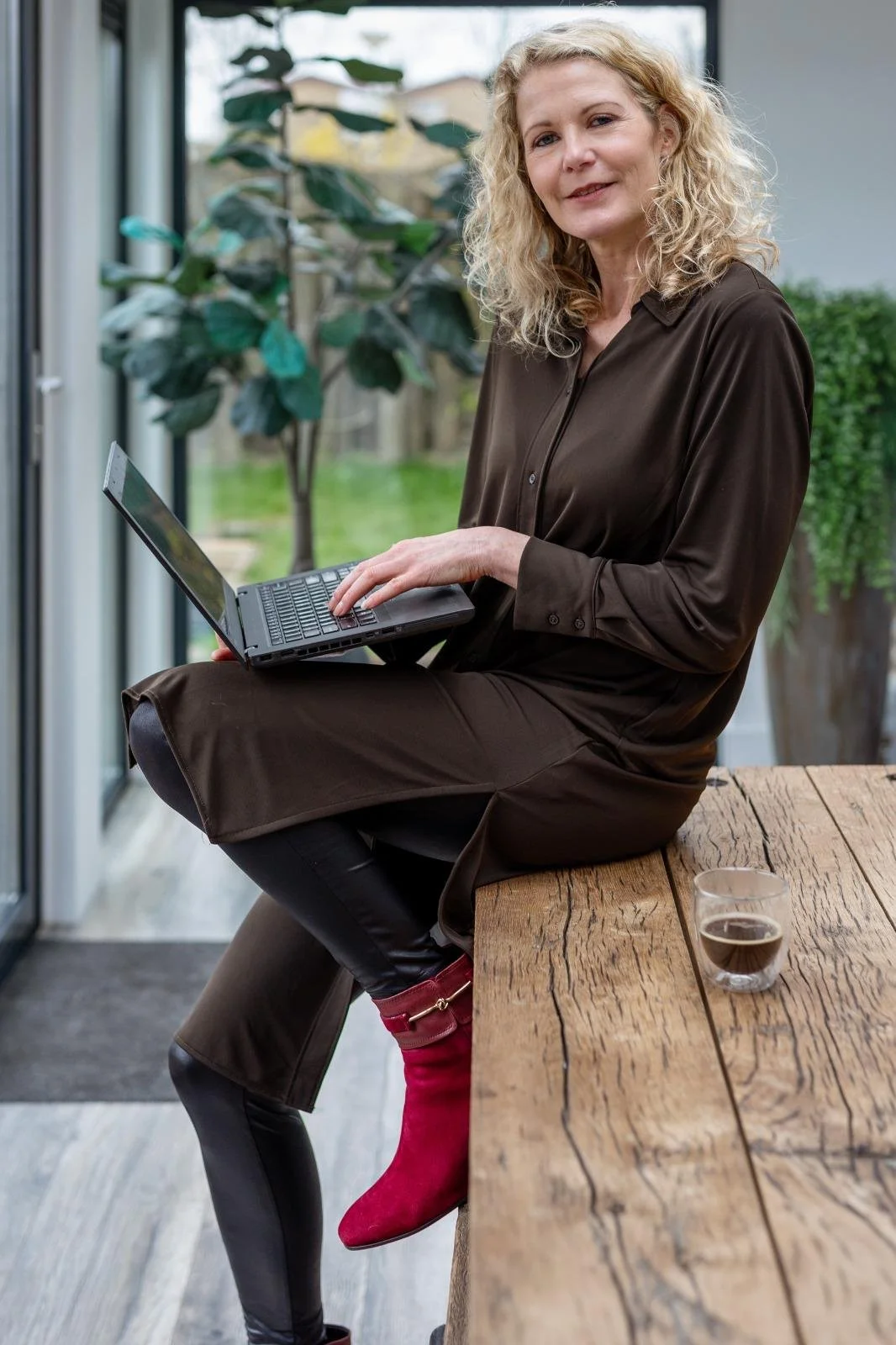Vrouw met krullend blond haar zit op de rand van een tafel met een laptop op schoot in haar coachingpraktijk in dordrecht.