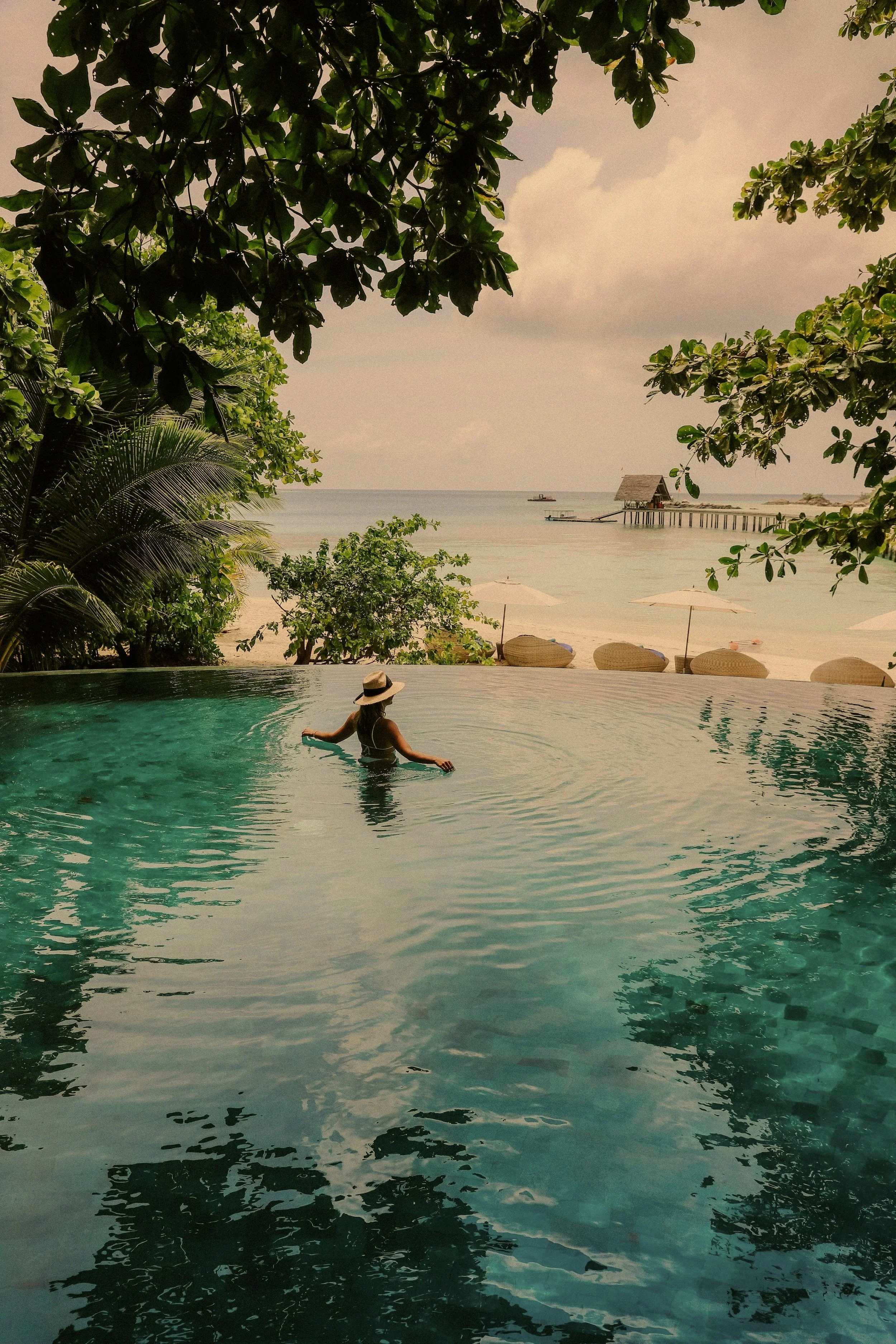 Person in a hat standing in an infinity pool with a view of the ocean, surrounded by lush greenery and a pier in the background.
