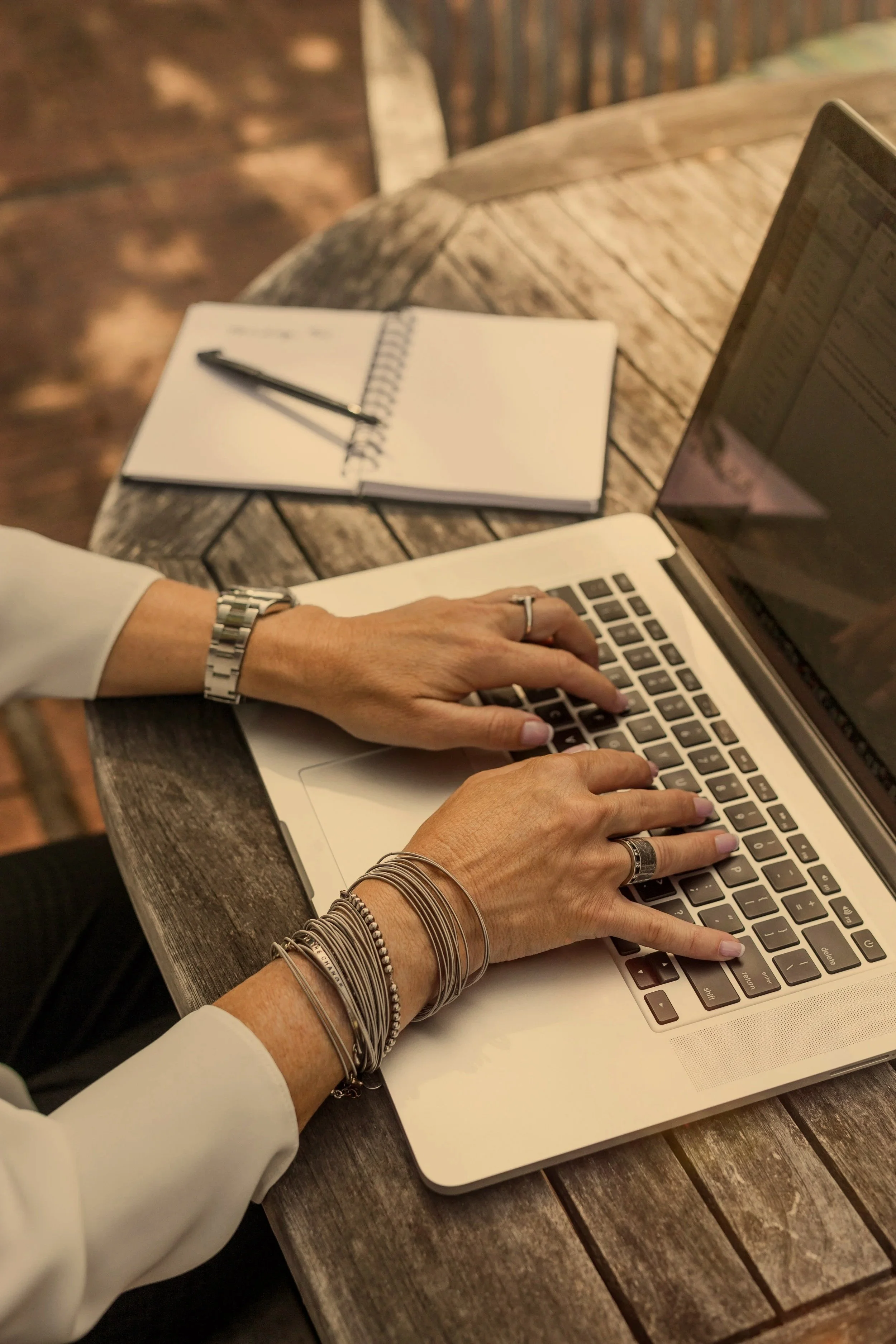 Person typing on a laptop at an outdoor wooden table, with a notebook and pen nearby.