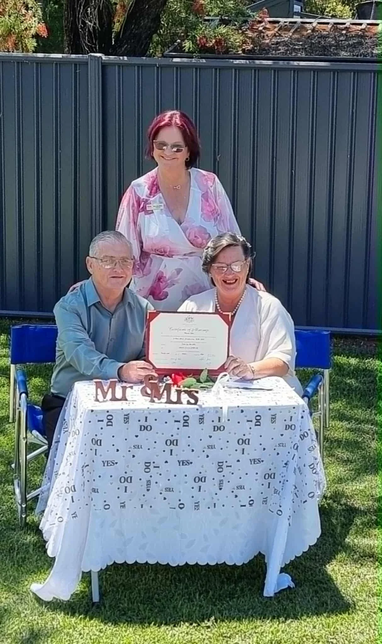 A couple at a wedding celebration sitting at a table outdoors, holding a wedding certificate, with a woman standing behind them. The table has a sign saying 'Mr & Mrs,' and the scene is sunny with a grassy background.