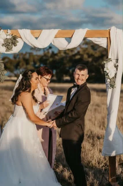 A bride and groom holding hands and smiling at each other during an outdoor wedding ceremony, with a woman officiant reading from a book under a decorated wooden arch in a field.