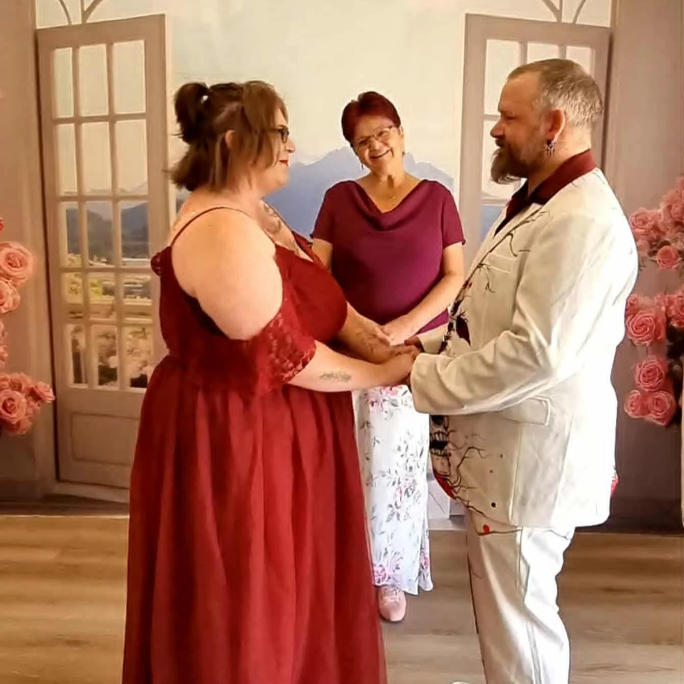 A couple getting married, holding hands, with a woman in a purple top officiating, in front of mountain landscape backdrop, surrounded by pink roses.