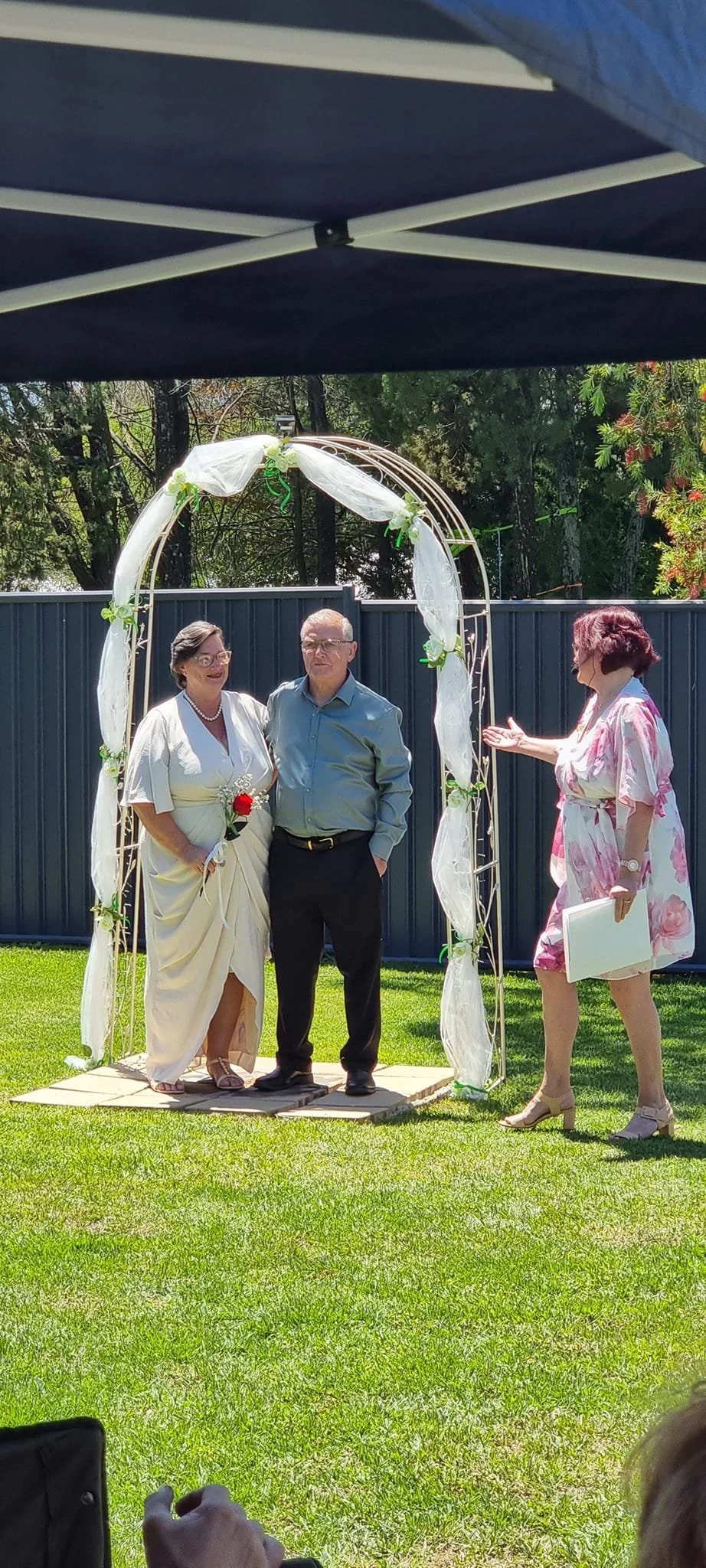 People exchanging vows at a wedding ceremony outdoors, with a decorated arch and a grassy lawn.