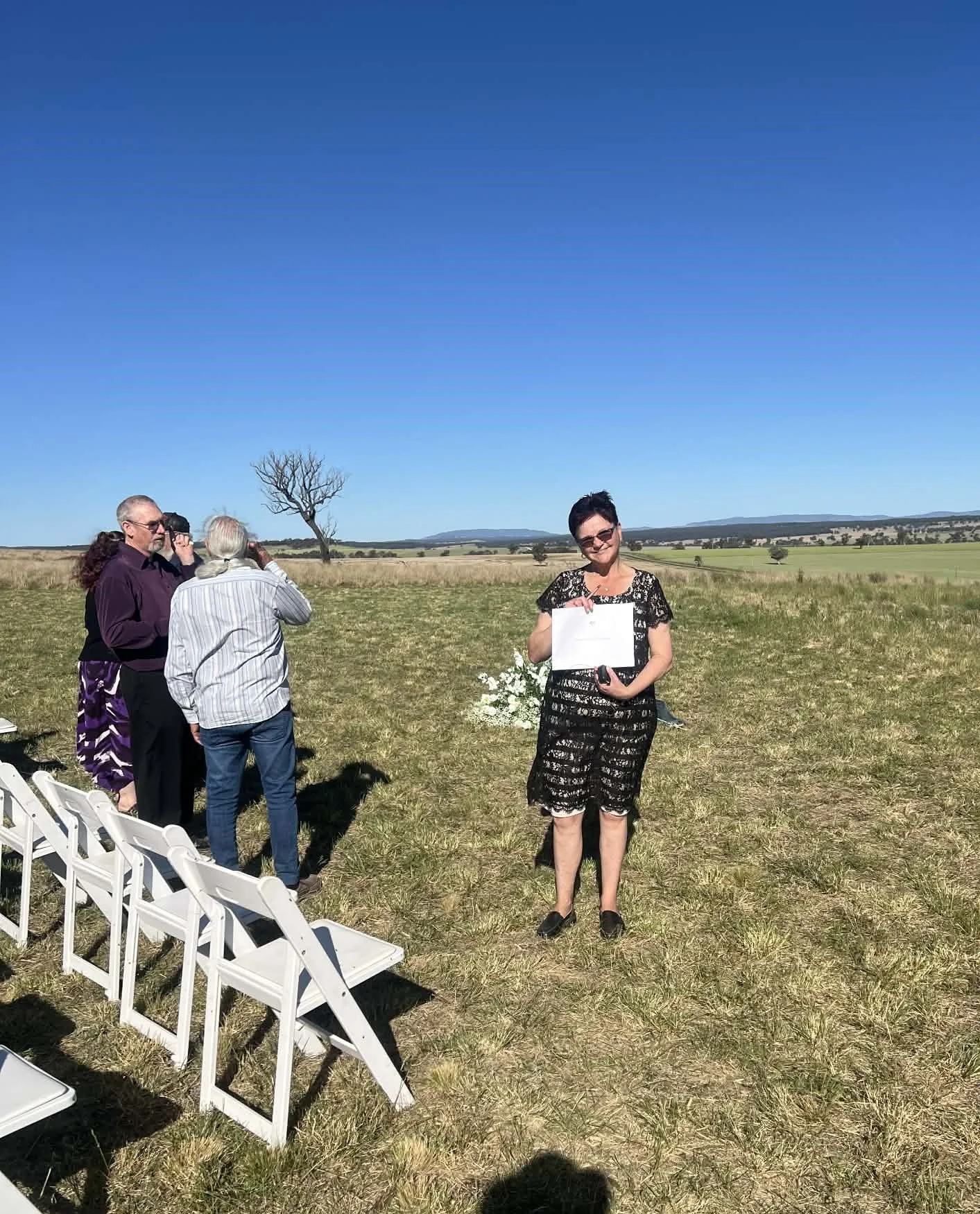 A woman standing in an open field holding a piece of paper, with three people behind her taking photos, and rows of white chairs in the grass on a sunny day.