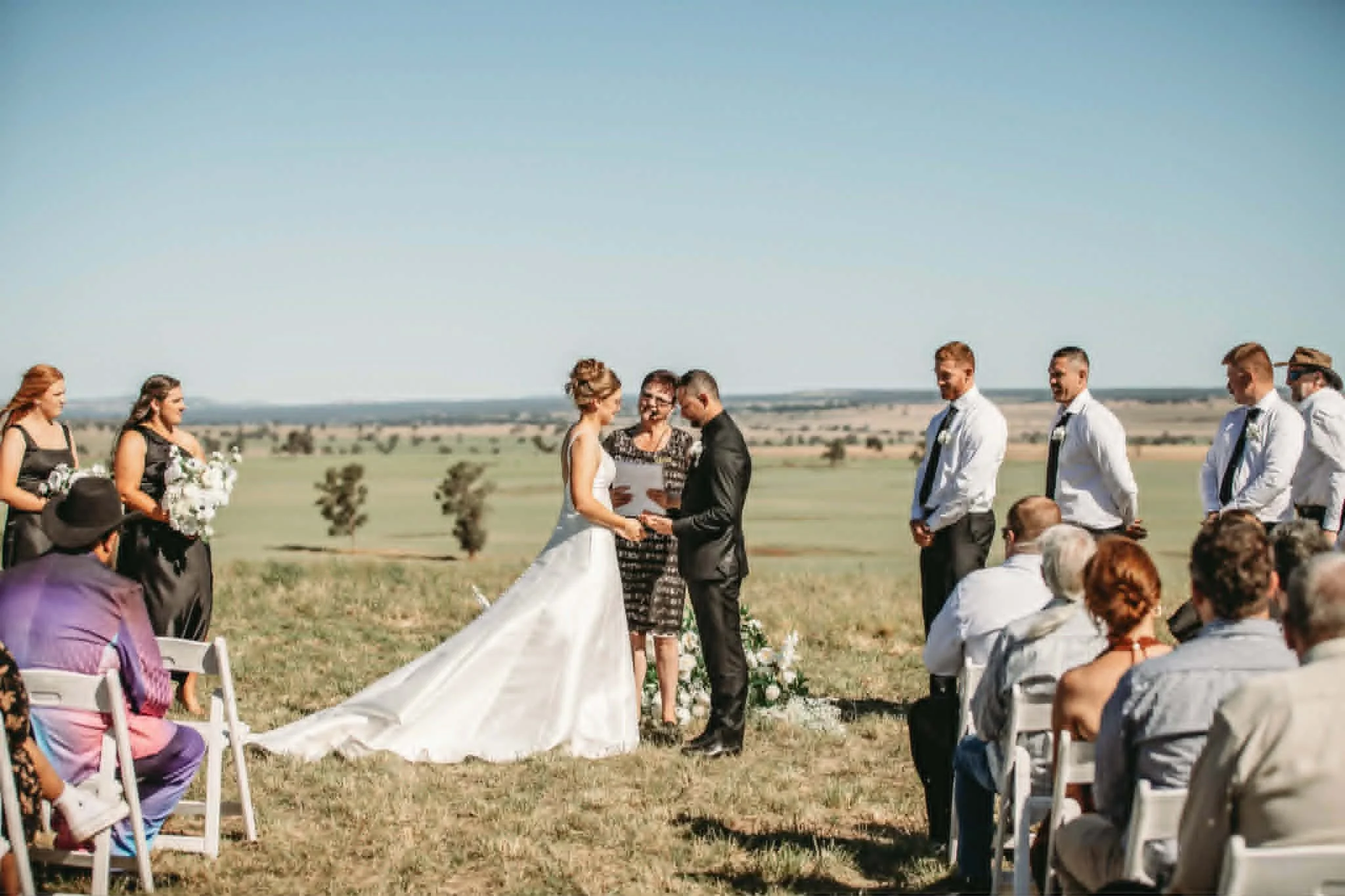 A couple getting married outdoors, exchanging vows in a field with onlookers and bridesmaids in black dresses holding floral bouquets, under a clear blue sky.