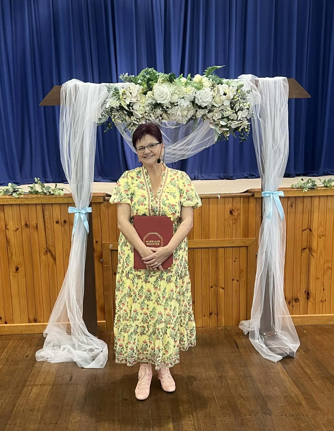 A woman stands in front of a wedding arch decorated with white flowers and tulle, holding a marriage register, in a venue with blue curtains and wooden paneling.