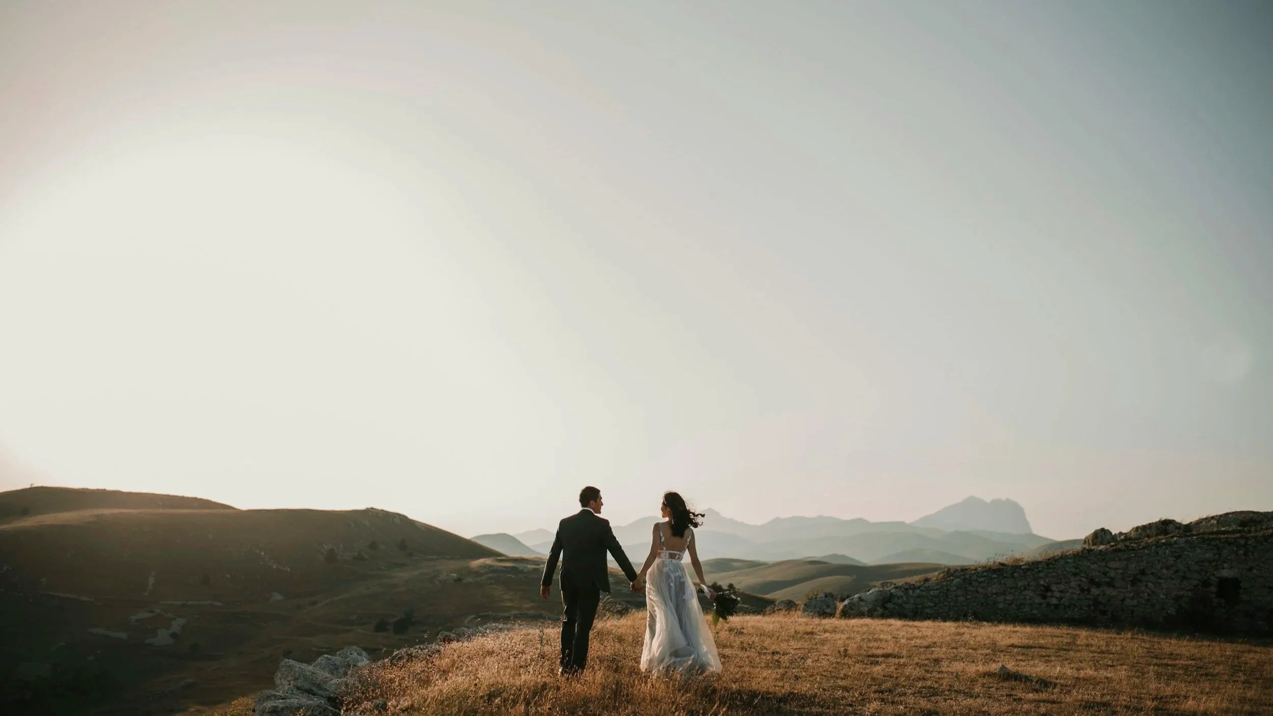 A bride and groom walking hand in hand in a grassy field during sunset, surrounded by rolling hills and mountains in the distance.