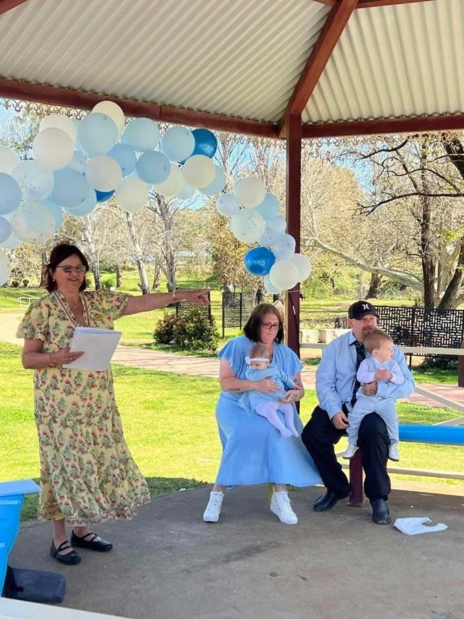 Family gathering under a gazebo with blue and white balloons, woman in floral dress speaking, three people sitting in casual attire, two children, outdoors in a park.