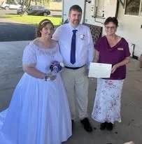 A bride in a white wedding dress and a groom in a light-colored suit with a blue tie, standing outside with a woman holding a certificate, possibly during a wedding ceremony or celebration.