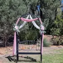 A decorated outdoor arch with pink and white ribbons and flowers over a park bench, trees and a fence in the background.