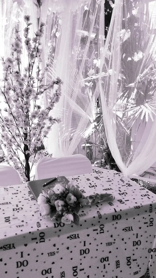 A decorated table with a bouquet of roses, a pen, and a photo, set against a backdrop of sheer curtains and flowering plants.