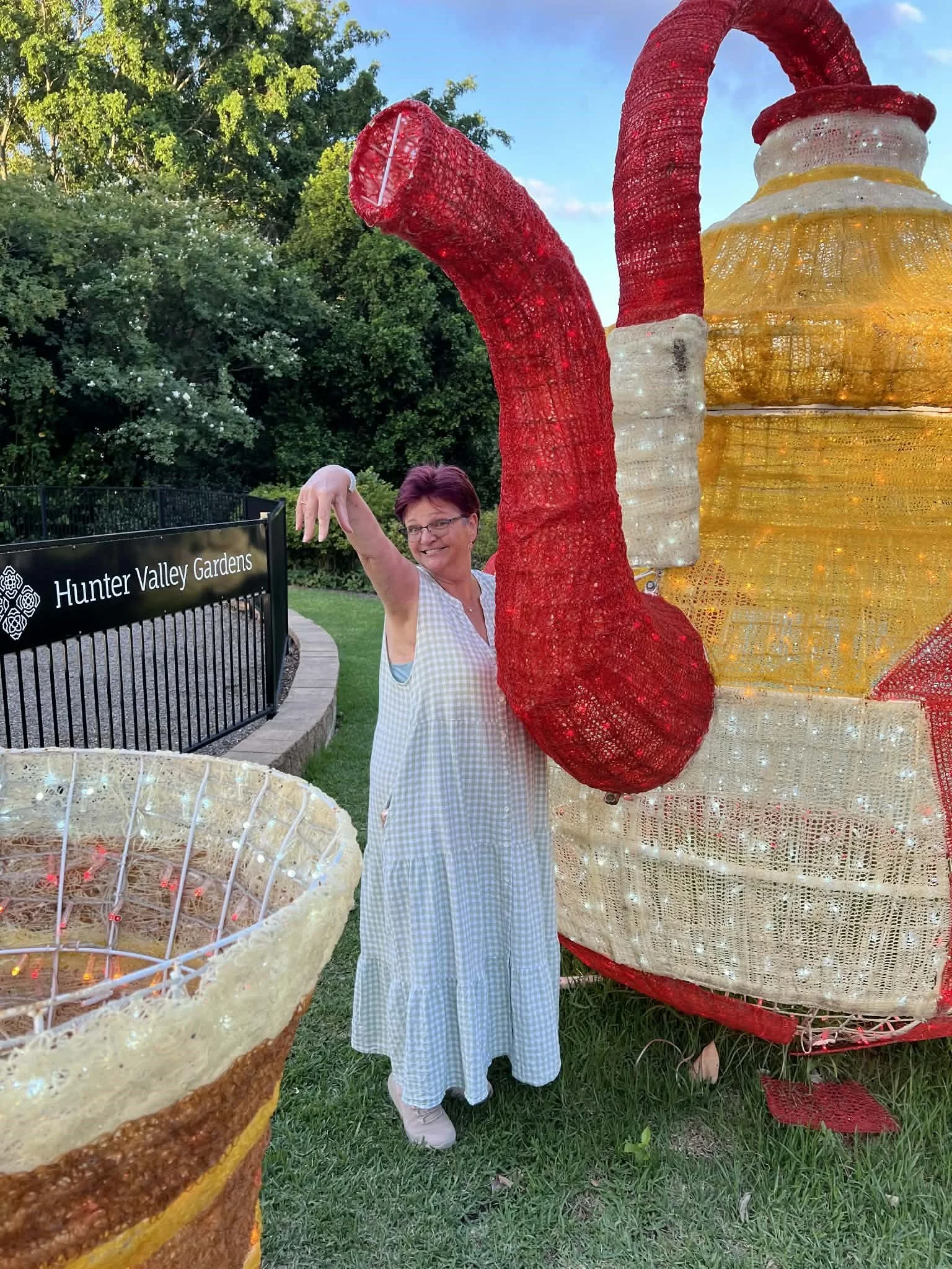A woman in a checkered dress smiling and posing near large illuminated holiday decorations resembling a Christmas tree and oversized ornaments, with a sign that reads 'Hunter Valley Gardens' in the background.