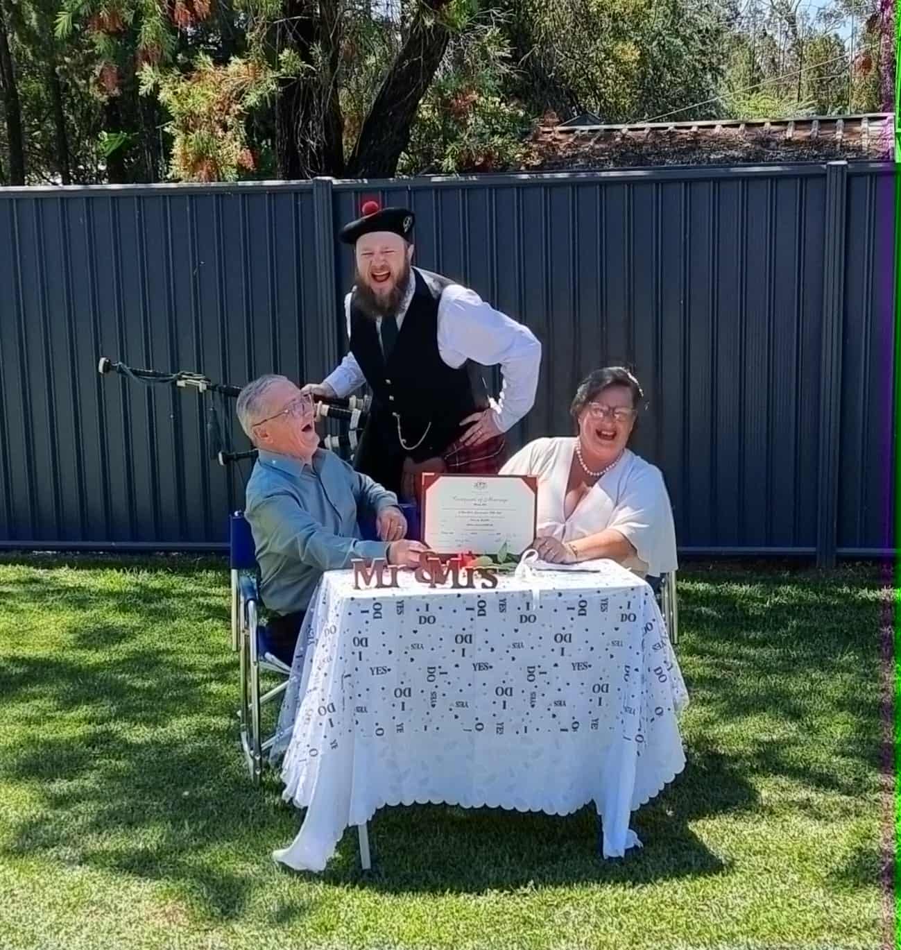 An elderly man with white hair and glasses and an elderly woman with short gray hair seated at a table outside, celebrating a wedding or anniversary with a framed certificate and decorative "Mr & Mrs" sign. A man dressed in Scottish traditional attire, including a hat with a red pom-pom, stands behind them, smiling and leaning forward in a festive manner. The scene is outdoors with a grass lawn, a black metal fence, trees, and a roof in the background.