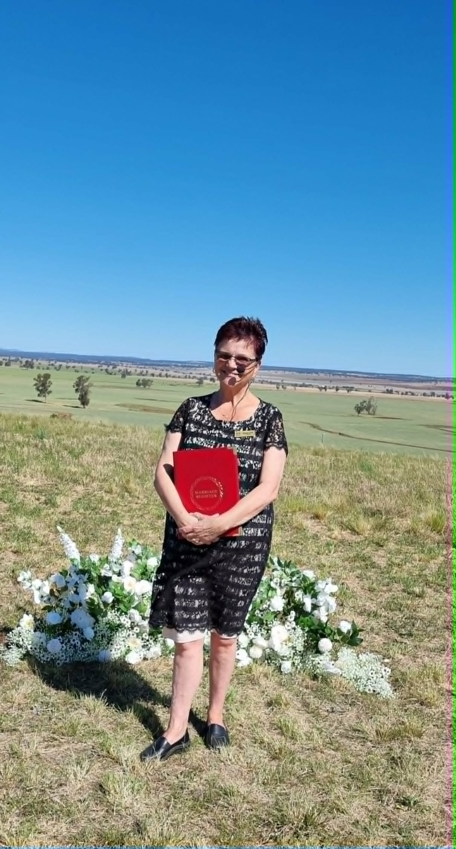 Woman in black lace dress holding a red folder, standing outdoors on a grassy hill with white floral arrangements and a rural landscape in the background.