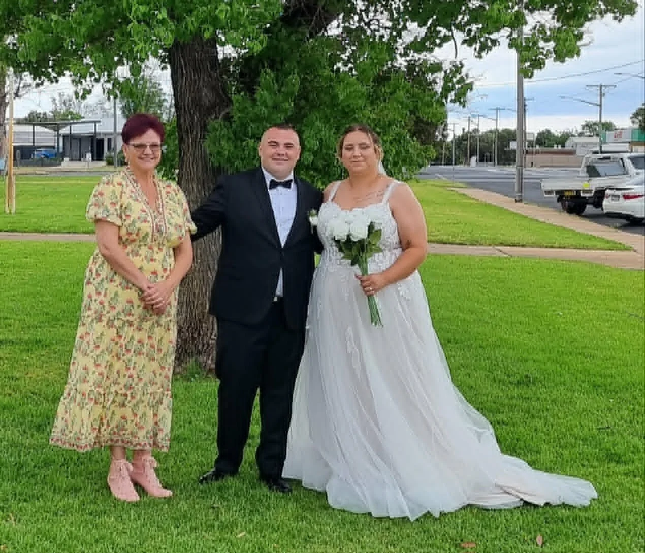 A bride in a white wedding gown holding a bouquet of white flowers, a groom in a black tuxedo, and a woman in a floral dress standing outdoors under a large tree with a grassy area and a street with cars in the background.