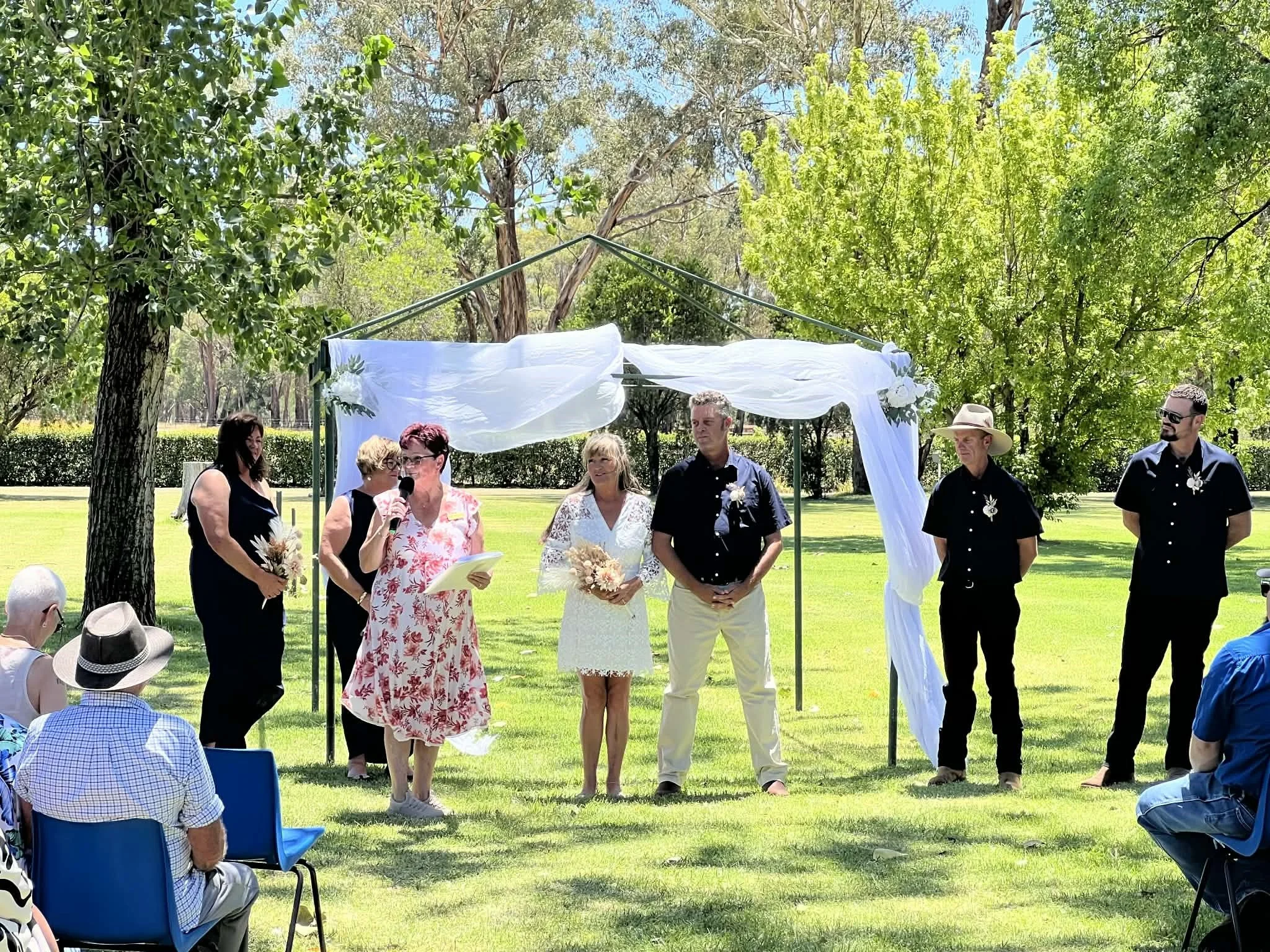 Outdoor wedding ceremony with a bride, groom, officiant, and wedding party standing under a decorated arch, with seated guests on grassy area surrounded by trees on a sunny day.