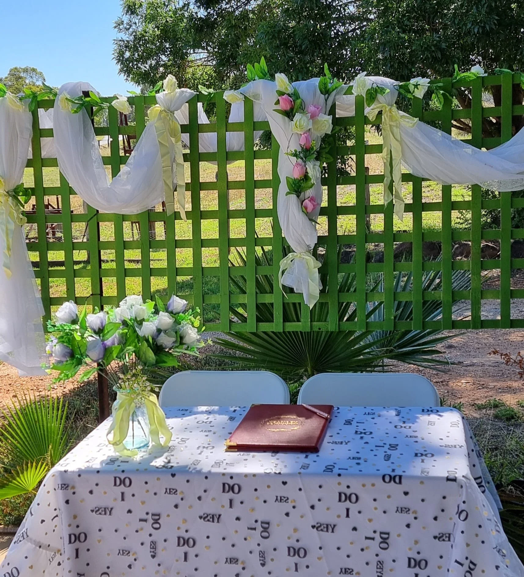 Outdoor wedding setup with a table covered in a white cloth patterned with the words 'I DO' and small hearts, two white chairs, a bouquet in a clear vase, and a green lattice decorated with white and pink flowers, white fabric drapes, and lush trees in the background.