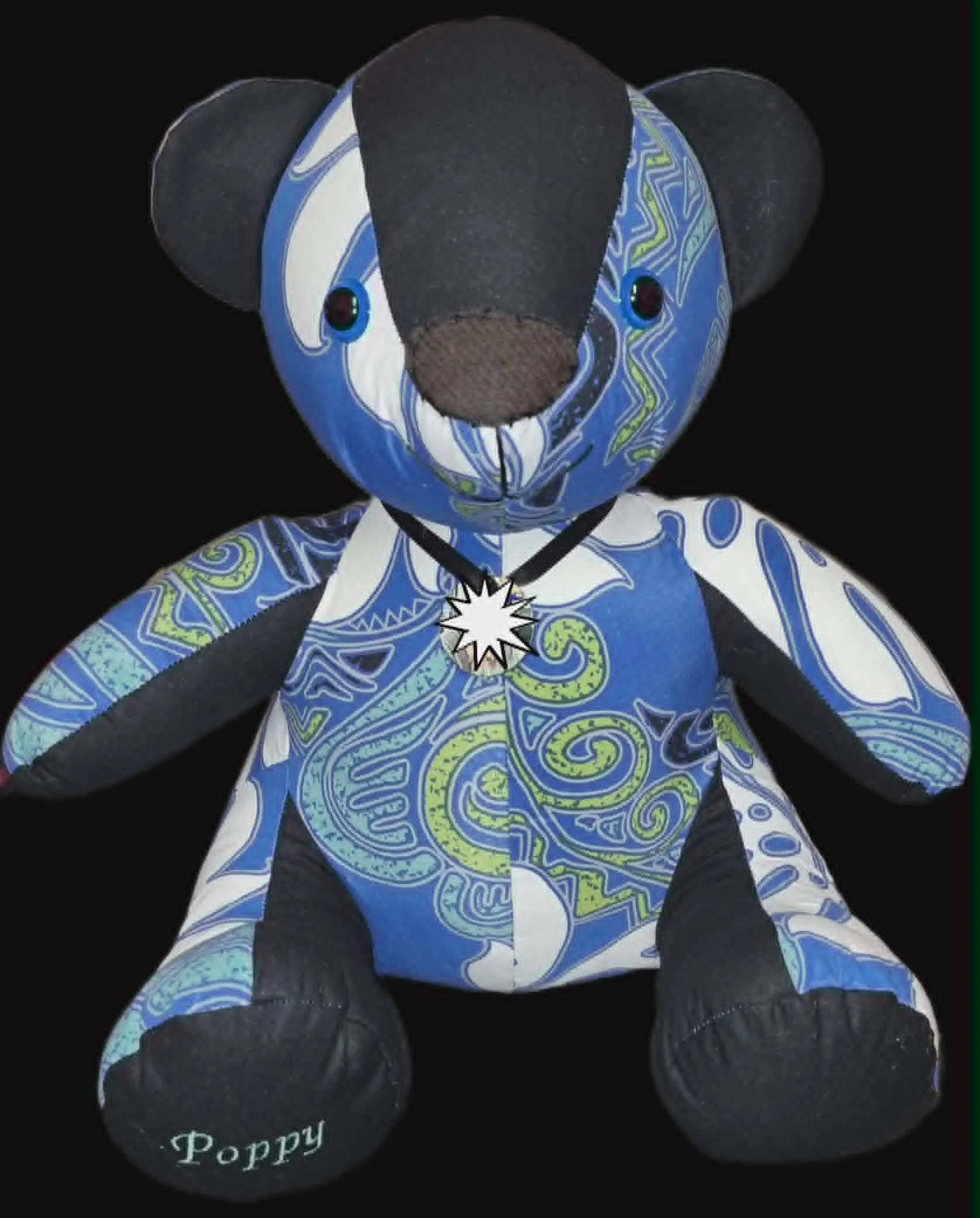 A stuffed teddy bear with black, blue, and patterned fabric, featuring a pendant necklace with a starburst design, sitting against a black background.