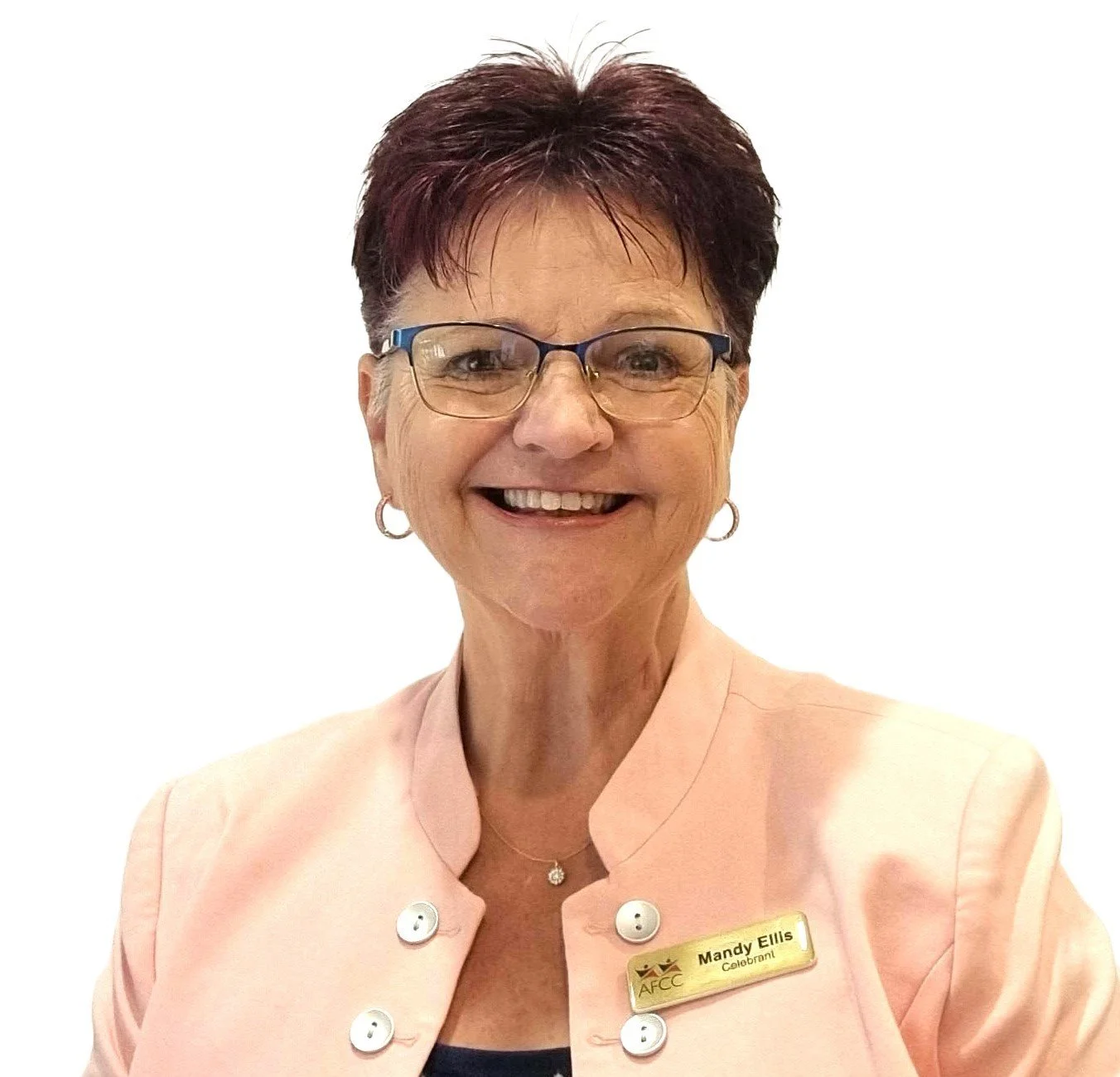 Portrait of Mandy Ellis, a woman with short dark hair, glasses, wearing a light pink blazer with white buttons and a name tag, smiling against a plain white background.