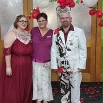 Three people standing in front of a Valentine's Day-themed backdrop with red heart decorations. They are dressed in semi-formal attire, smiling and posing for the photo.