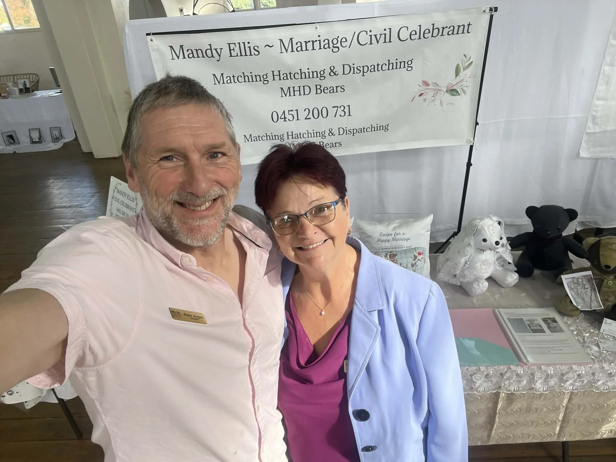 Two smiling people taking a selfie in front of a display table at a wedding celebration. The man has gray hair and a beard, wearing a light pink shirt with a name tag. The woman has short dark hair, glasses, and is wearing a purple top with a light blue jacket.