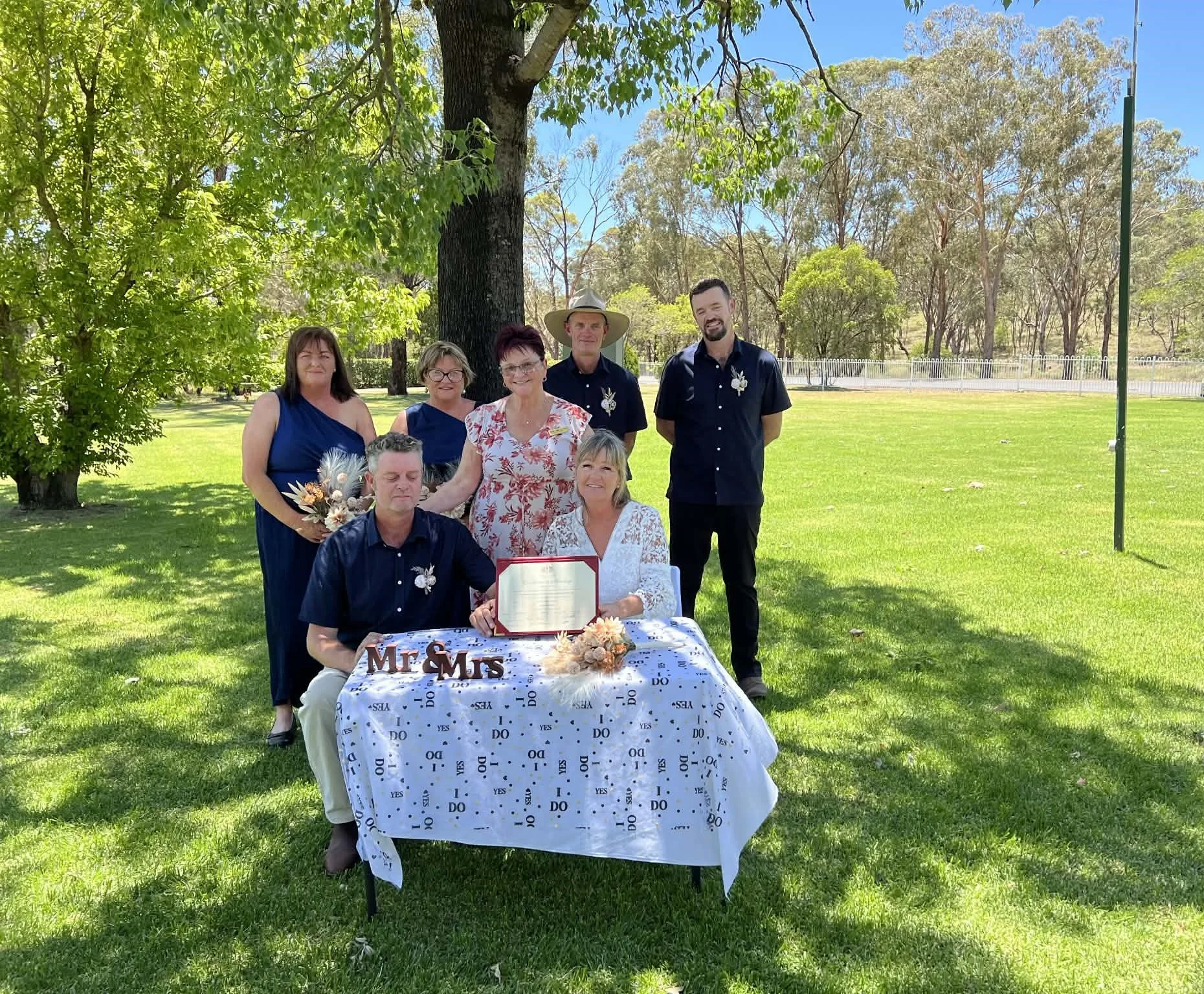 A group of seven people gathered outdoors under a large tree during a wedding celebration. They include a bride and groom sitting at a table with a 'Mr & Mrs' sign, holding a certificate, and five other guests standing behind them. The scene features bright sunlight, green grass, and clear skies.